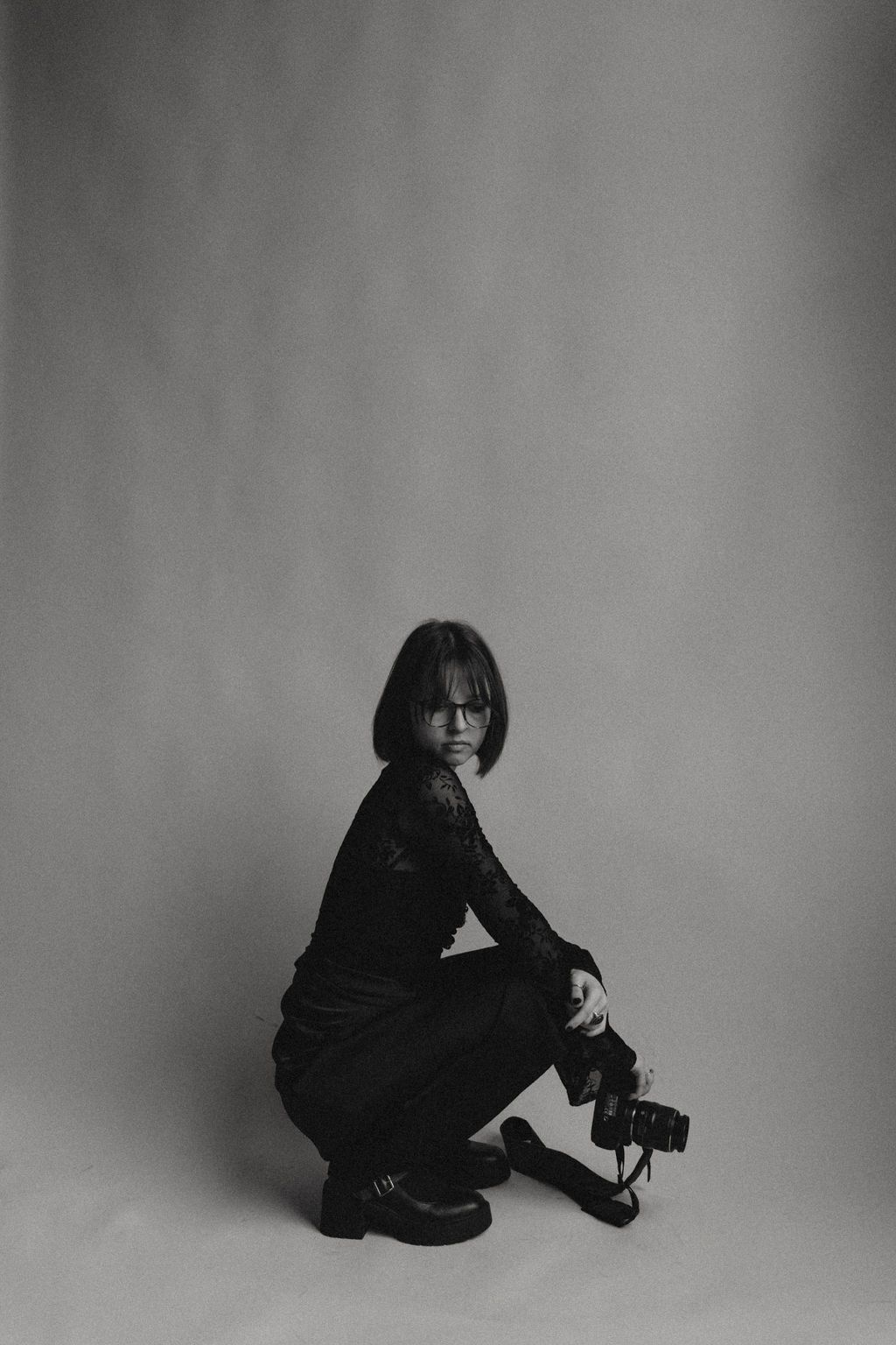 Black and white portrait of a photographer in dark clothing crouching with camera against plain backdrop.