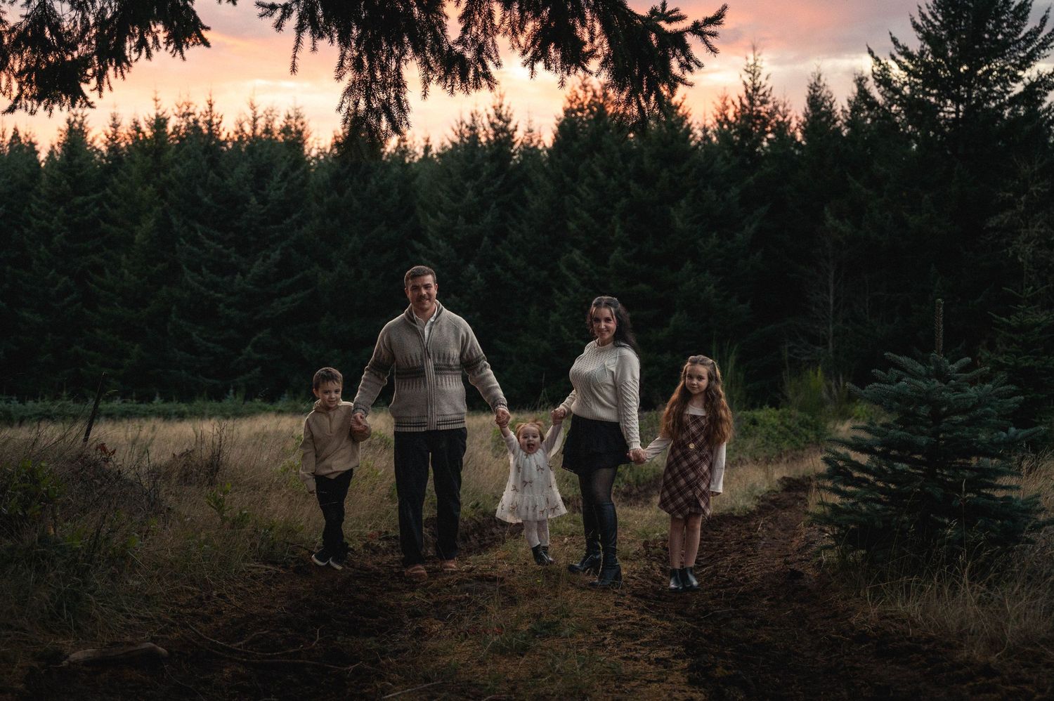 A group walks through an evergreen forest at sunset with warm pink skies overhead.