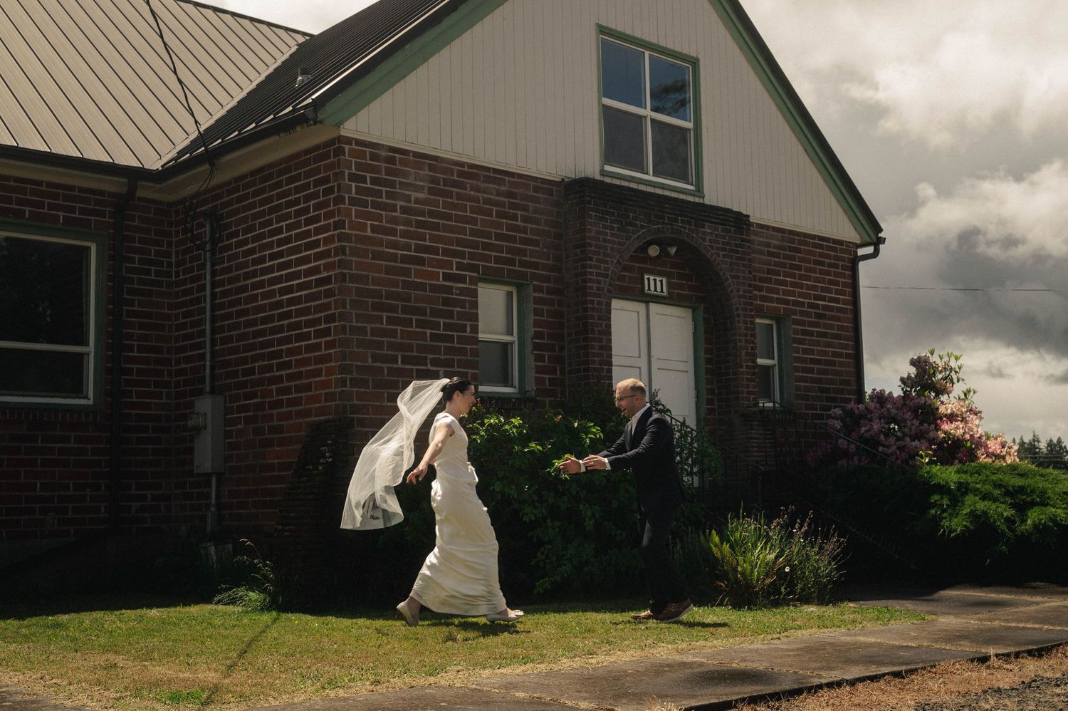 Bride and groom walking hand in hand outside a rustic brick church under dramatic stormy skies on their wedding day.