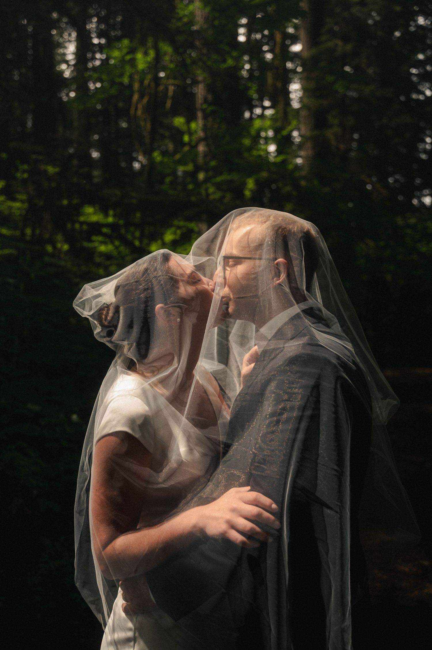 A bride and groom share a romantic kiss beneath a sheer wedding veil in a lush forest setting.