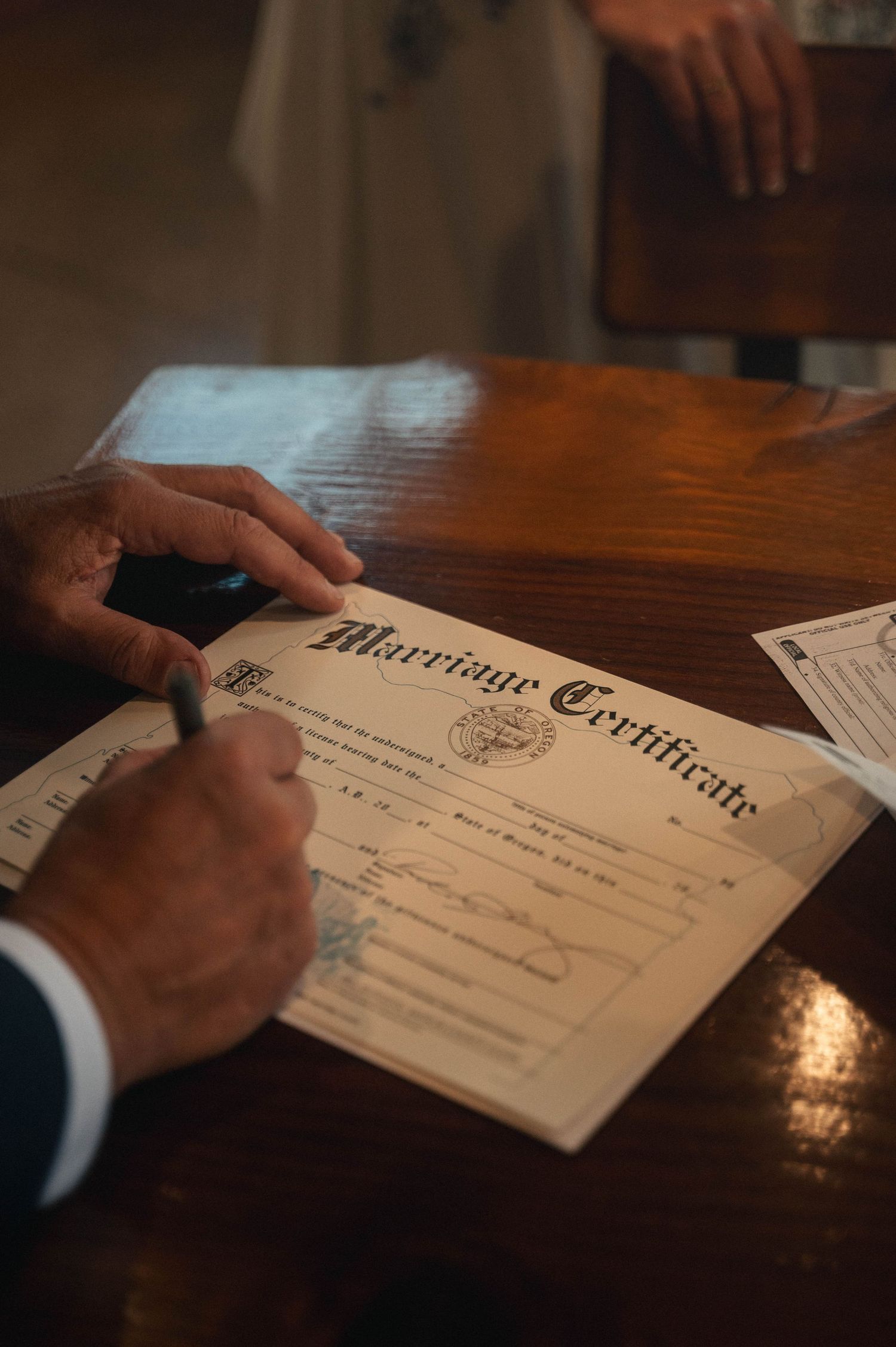 Person signing a marriage certificate on a wooden table, with another person standing nearby.