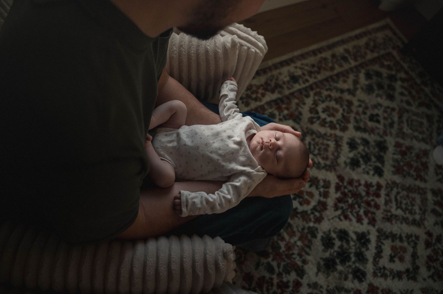 A baby lies on a patterned carpet while hands reach to adjust their white onesie.