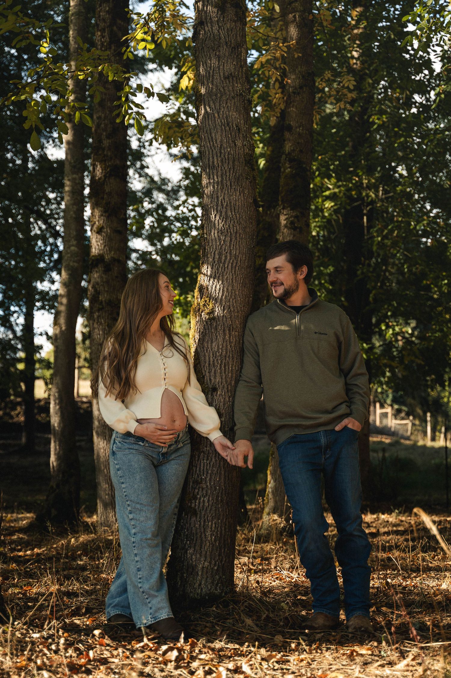 A couple stands together by a tree trunk in a wooded area during golden hour, wearing casual fall attire.