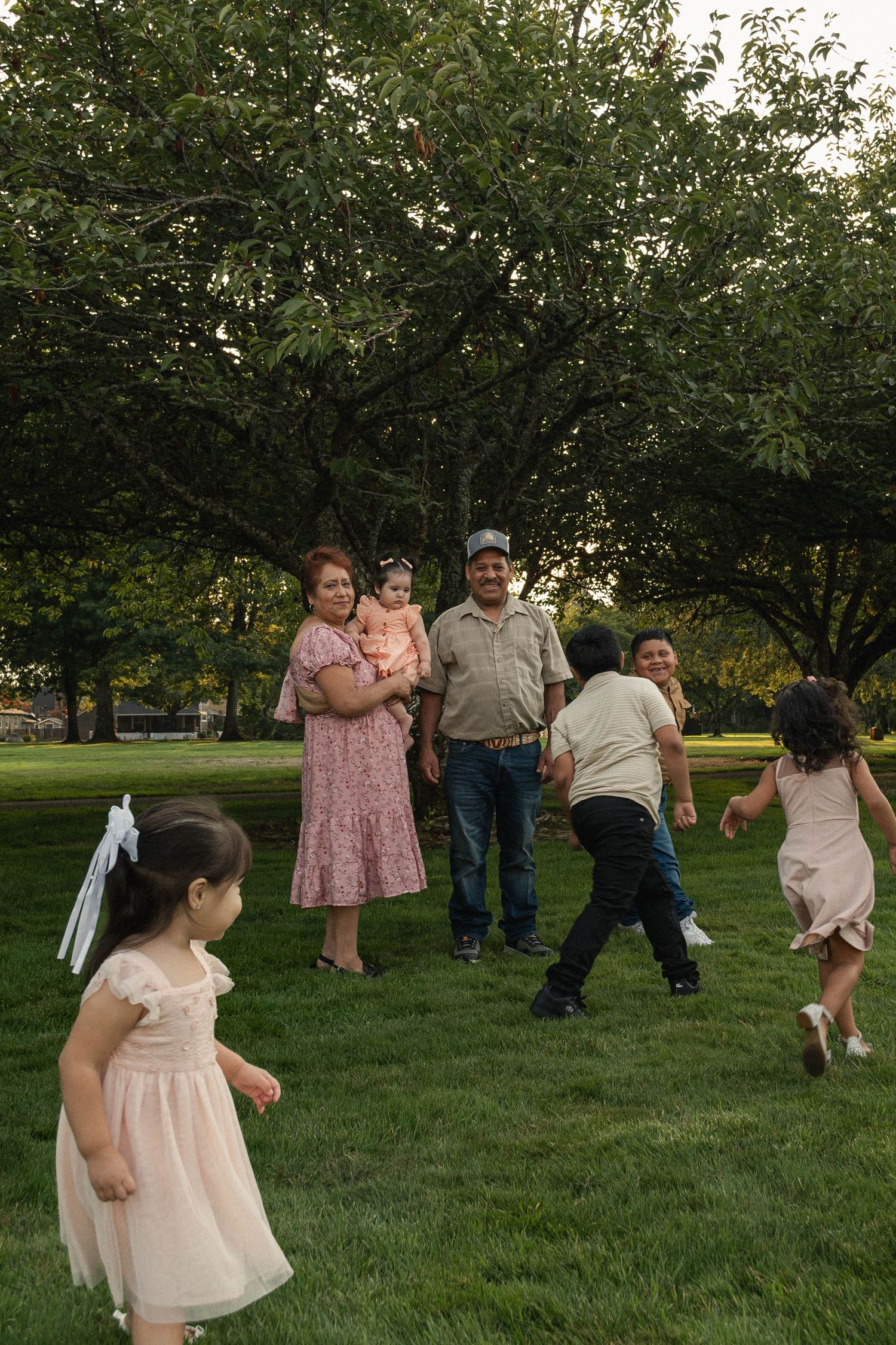 Grandparents holding their infant grandchild in a grassy park while 3 other grandkids run playfully around them.