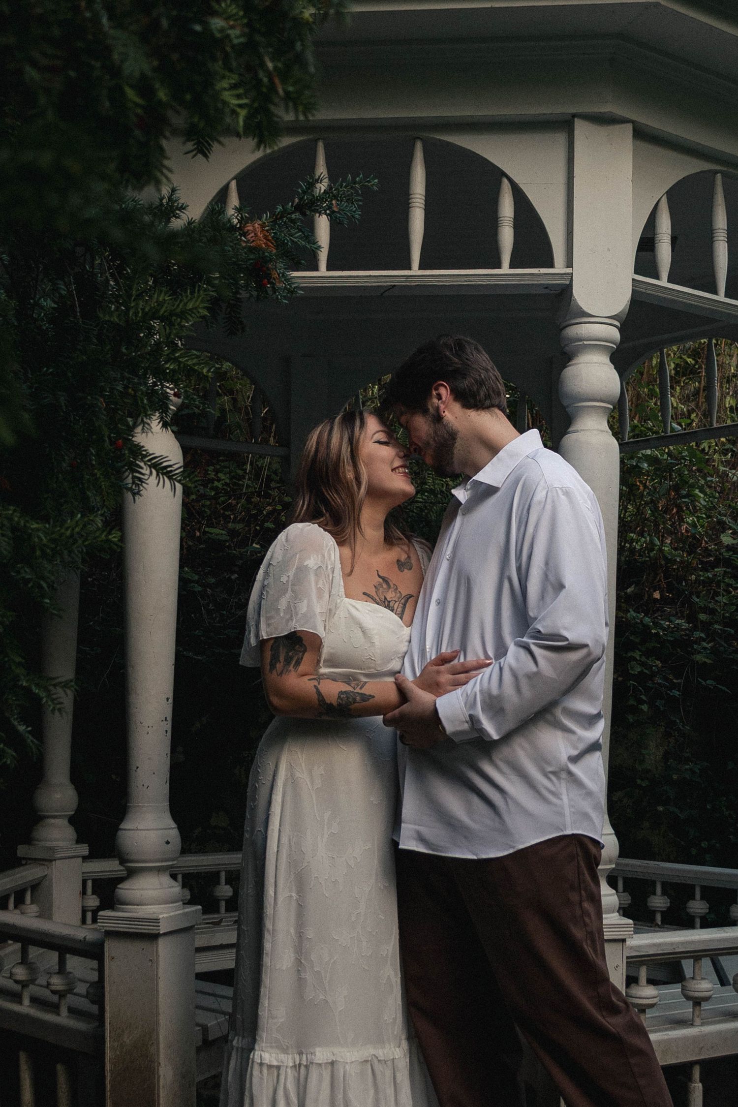 CIS couple standing close, holding hands and laughing under a pergola surrounded by ferns at Deepwood Museum in Salem Oregon.