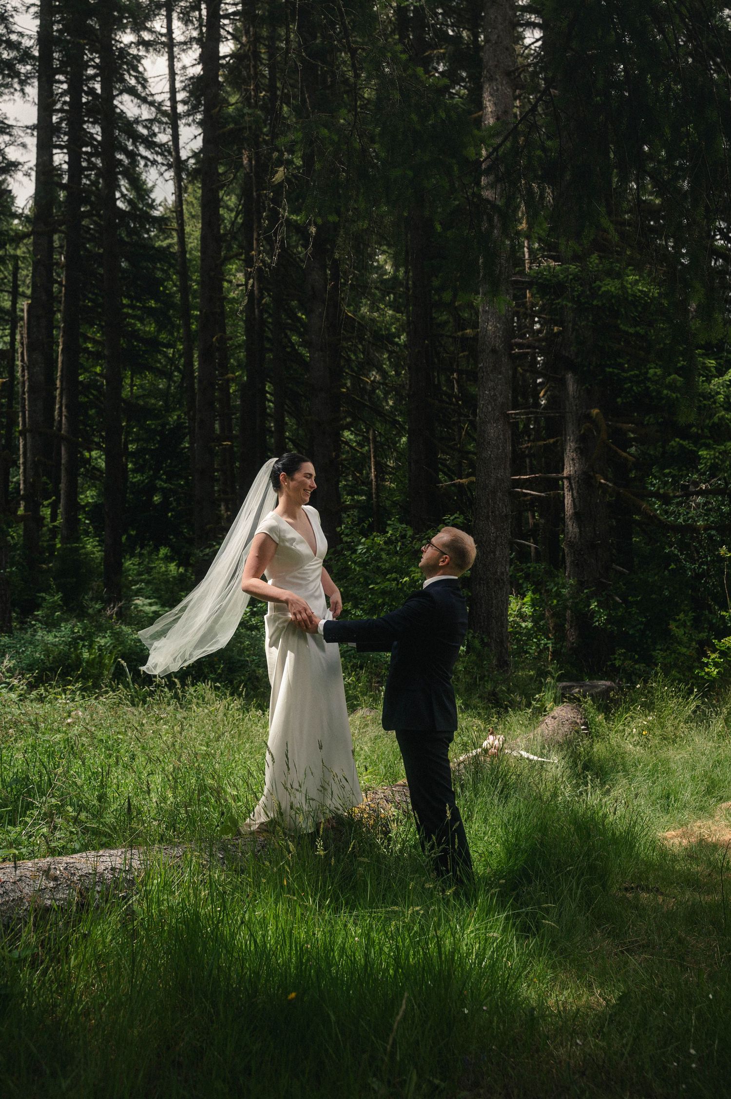 Bride and groom hold hands in a lush forest clearing, her veil flowing in the breeze during an outdoor wedding.