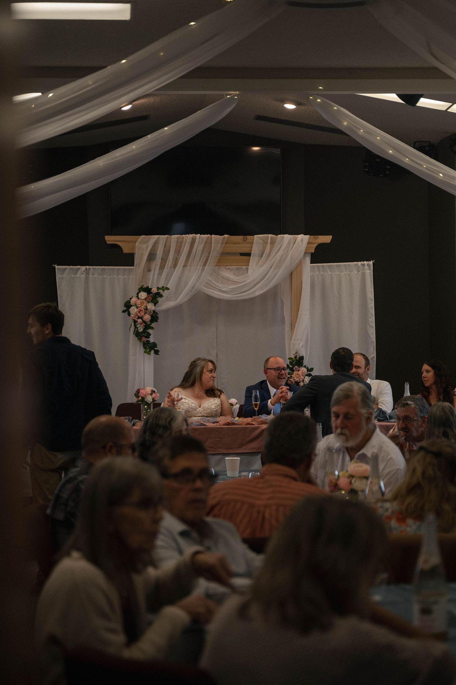 Dimly lit wedding reception with guests seated at tables under draped ceiling decor.
