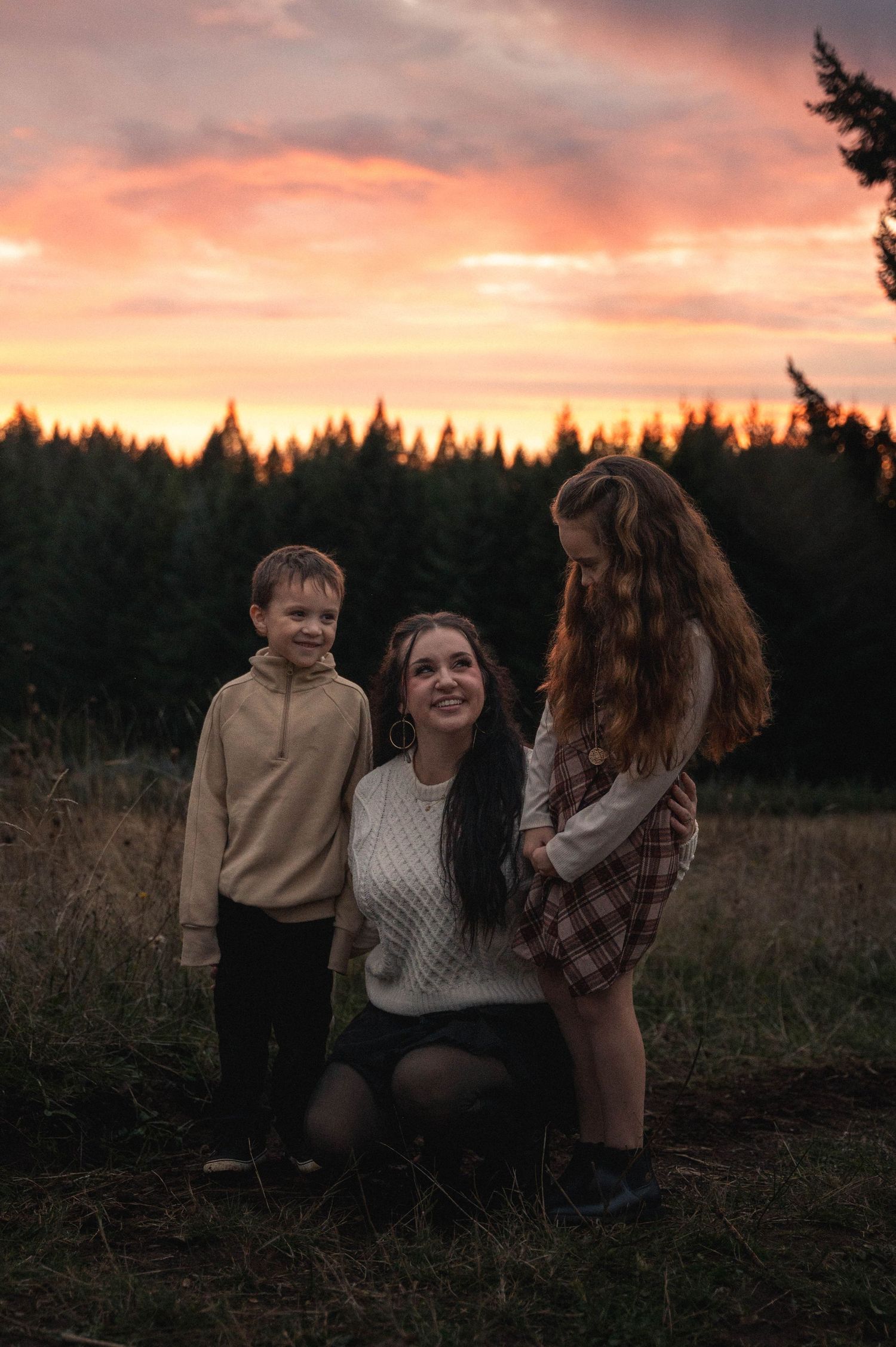 A family stands together in a grassy field at sunset with evergreen trees silhouetted against an orange and pink sky.