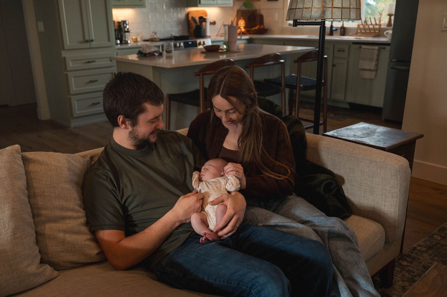 A couple sits together on a gray couch in a cozy living room with soft lighting and exposed brick walls.