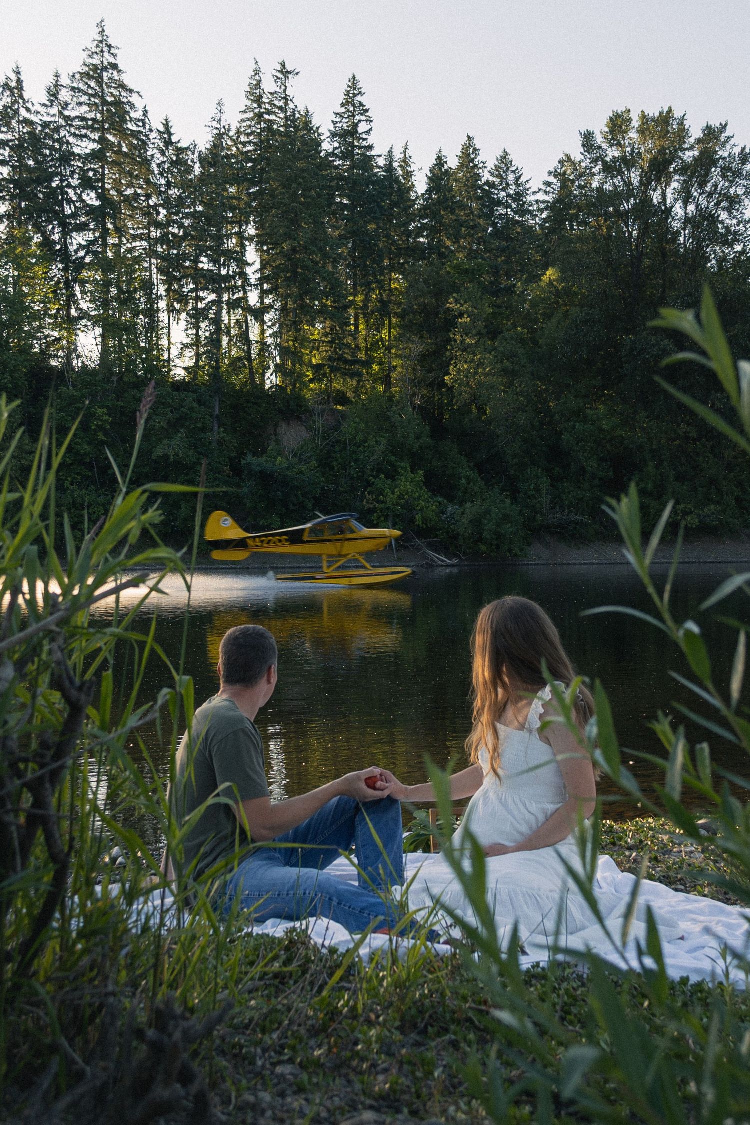 expecting couple sitting, holding hands by the river at Willamette Mission State Park while a water plane skips past
