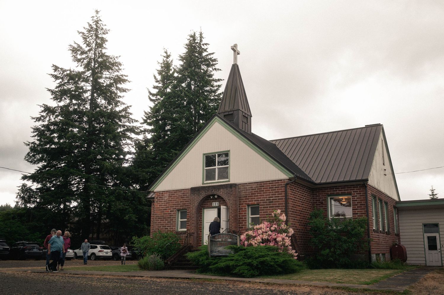 Small brick church with steeple cross, tall evergreen trees, and blooming pink shrubs under overcast sky.
