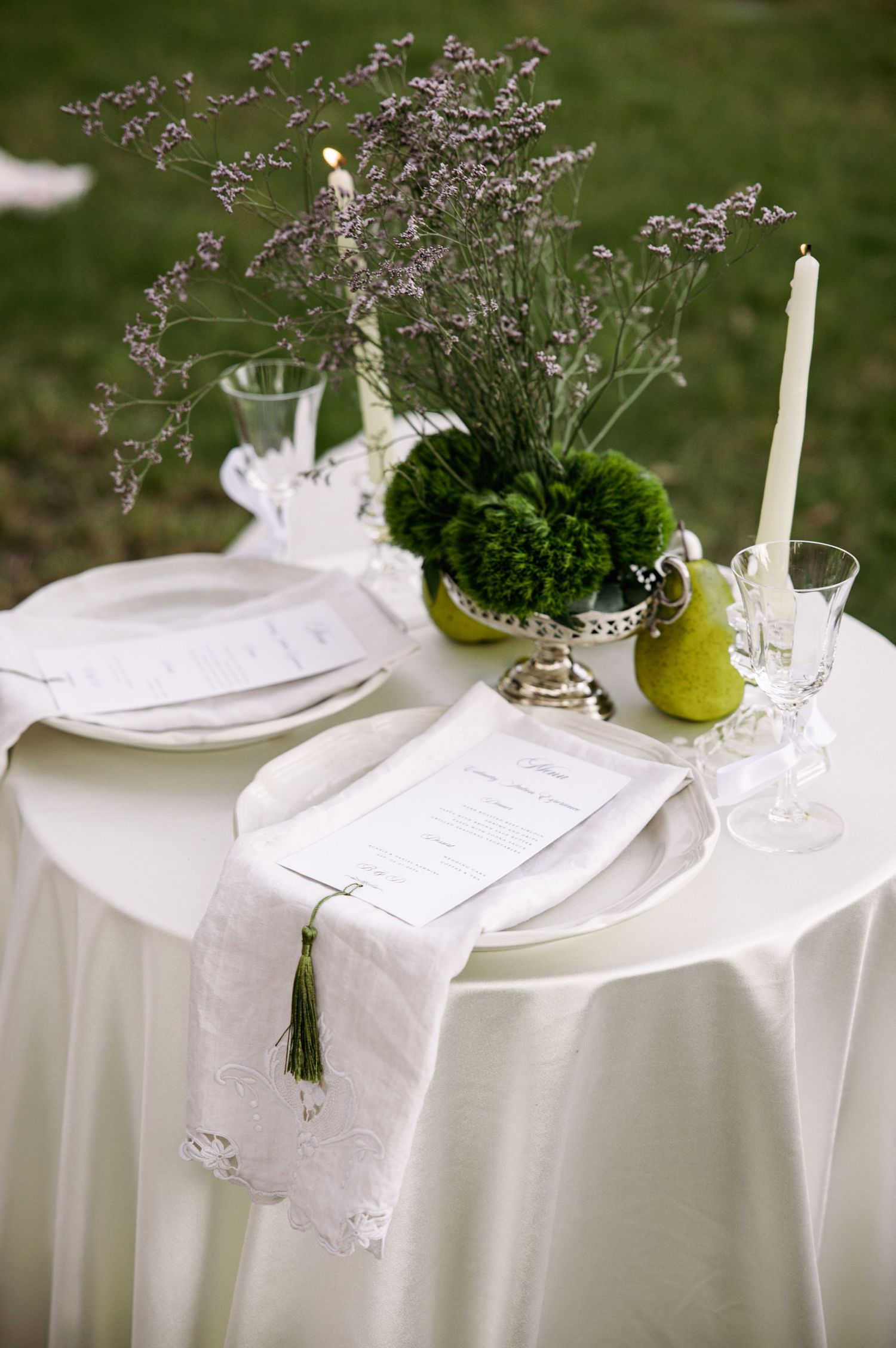 Elegant outdoor wedding table setting with white linens, green floral centerpiece, pears, lavender, and white taper candle.
