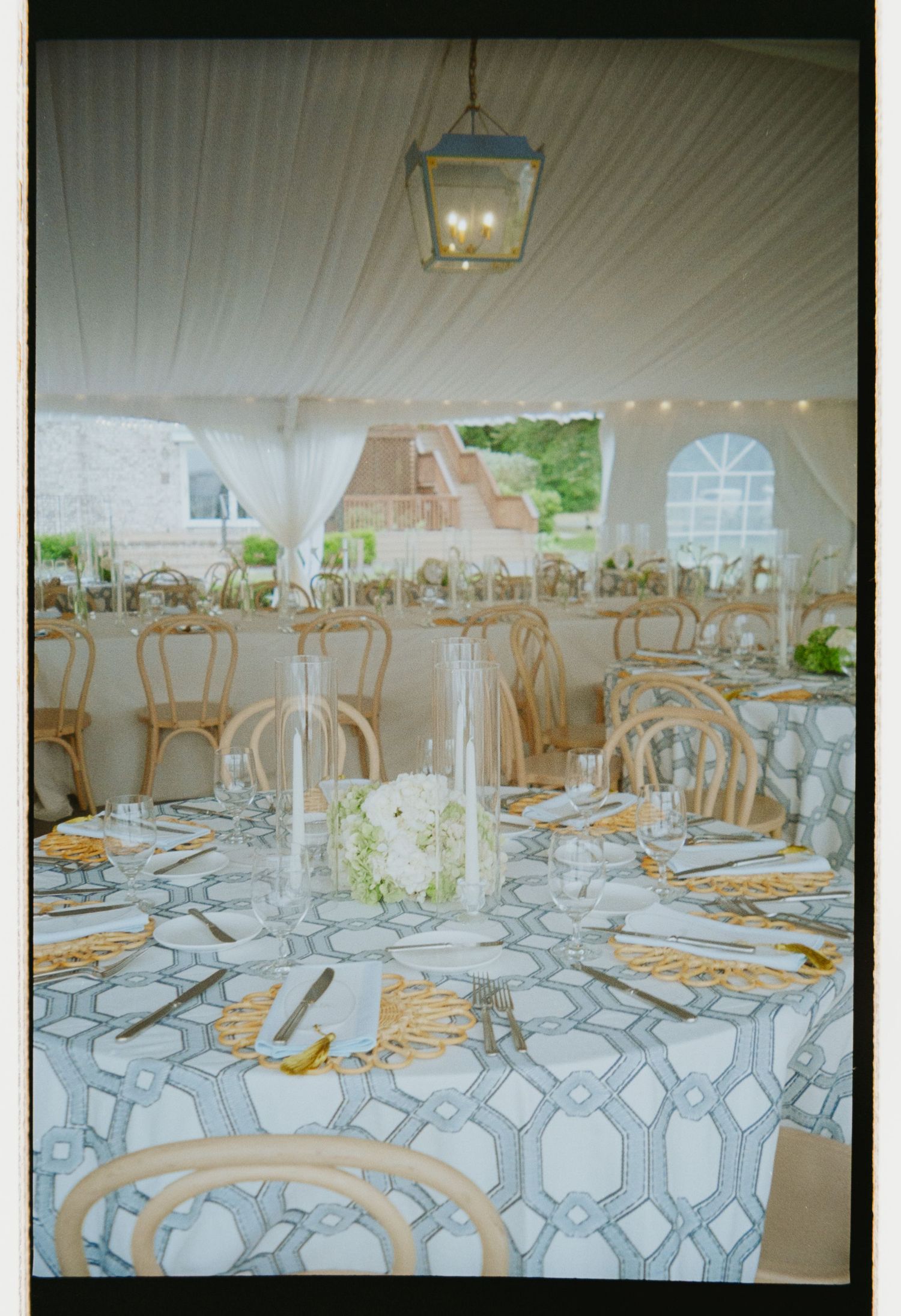 Elegant wedding reception tent with patterned tablecloths, gold chargers, floral centerpieces, and string lights.