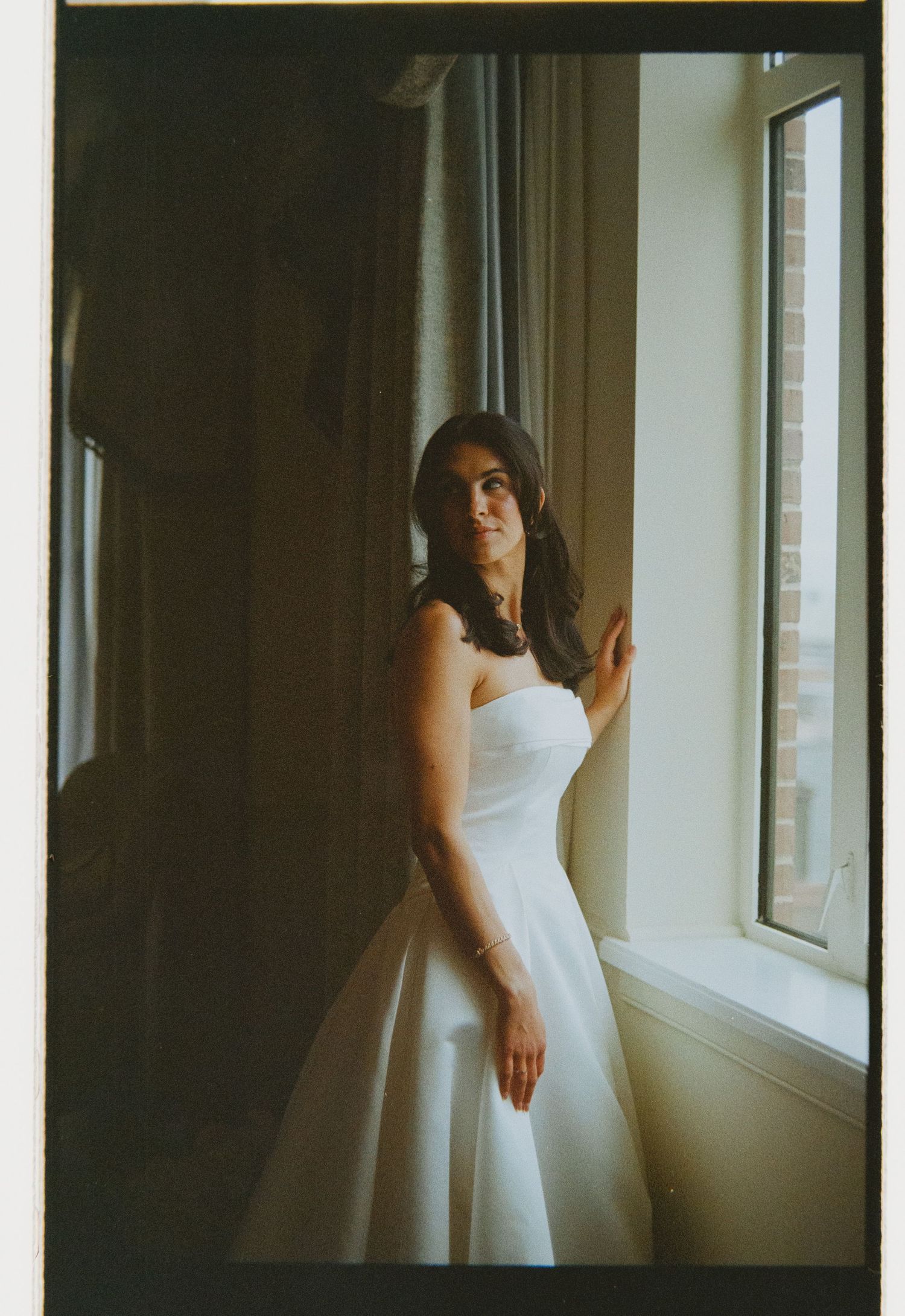 A bride in a white strapless ball gown stands by a sunlit window, gazing thoughtfully in a film photography style.