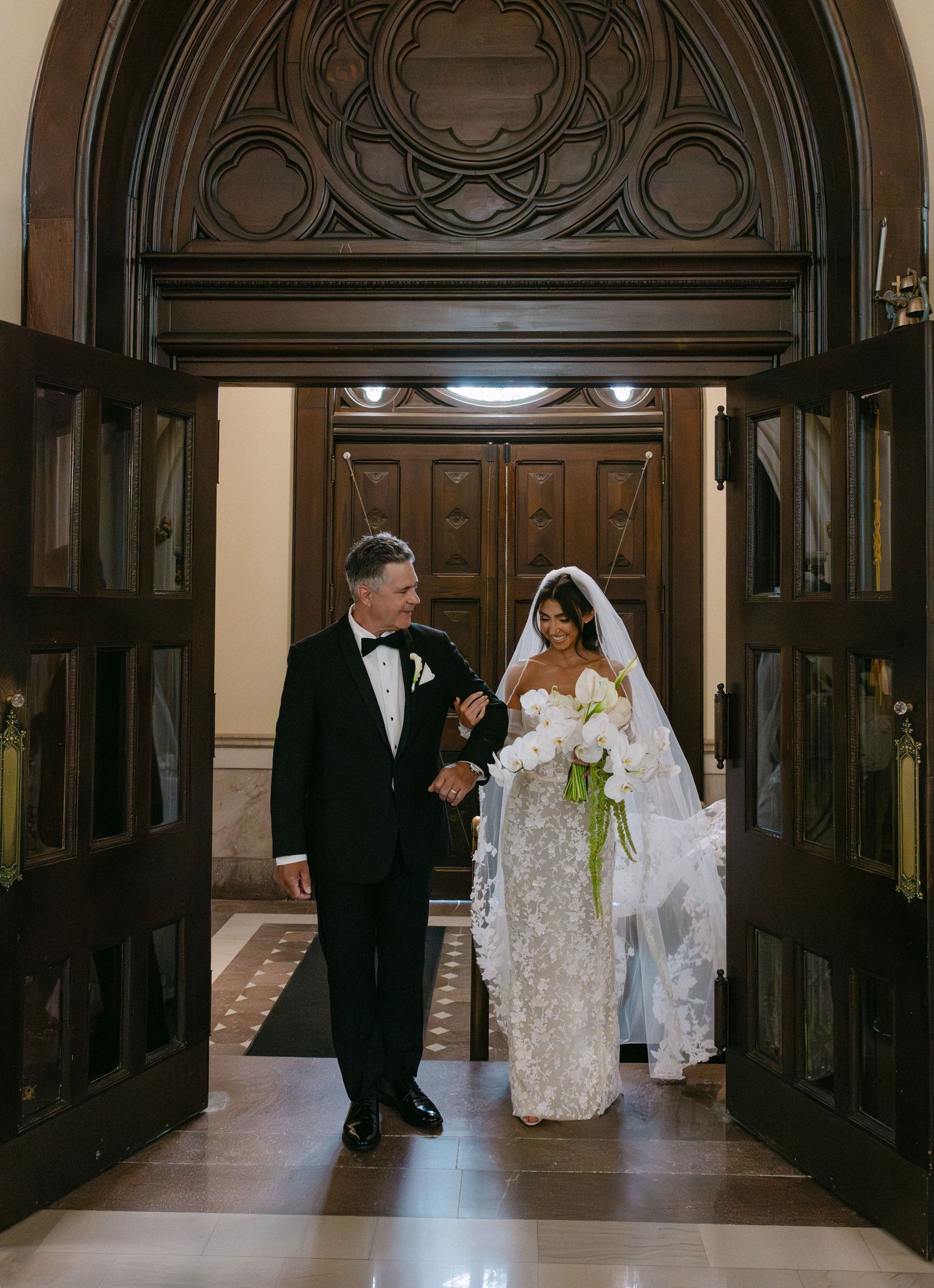 Wedding couple proceeds down a grand hallway with ornate wooden paneling and architectural details.