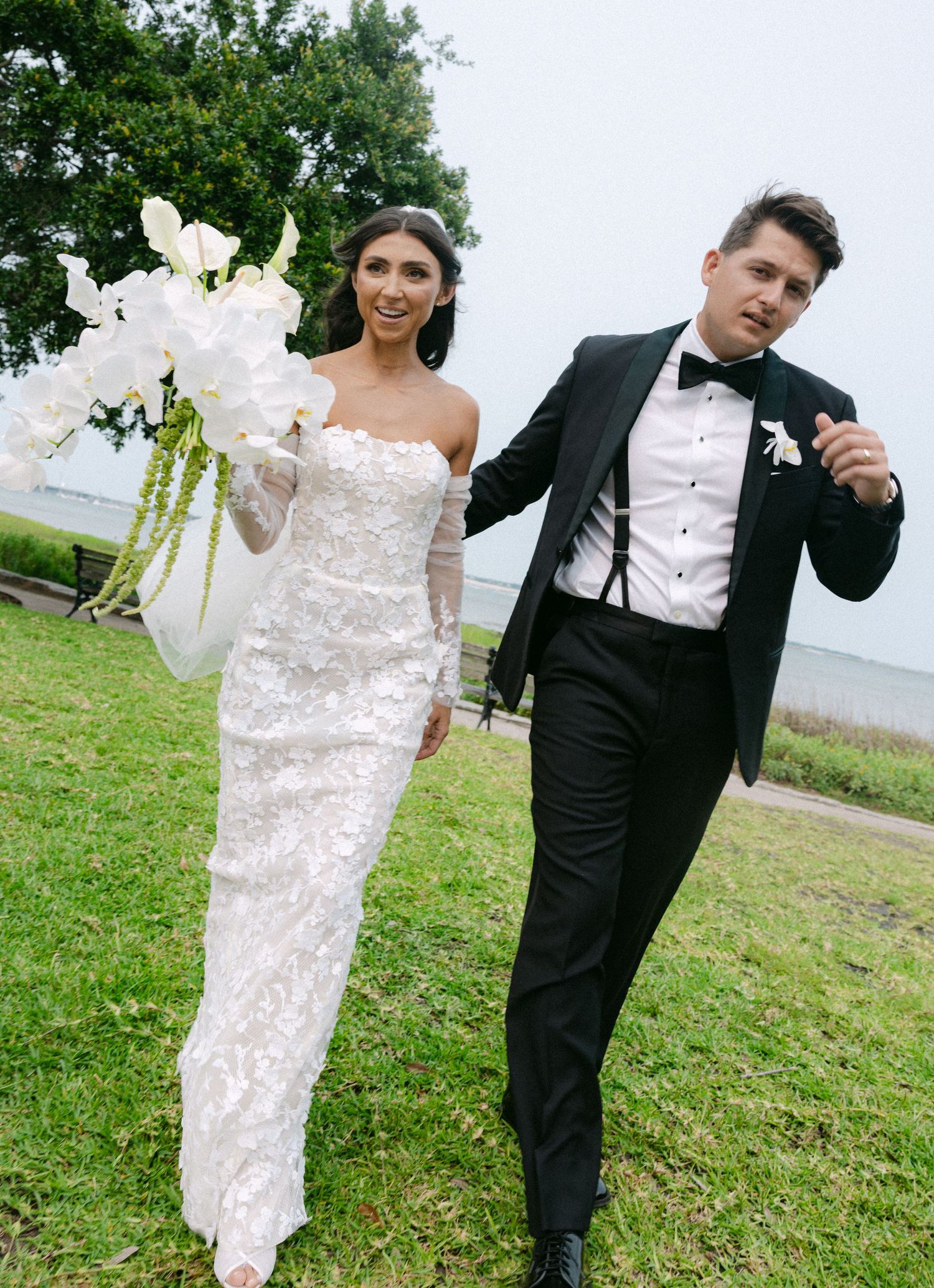 Wedding couple playfully poses on grassy field with white floral arrangement.