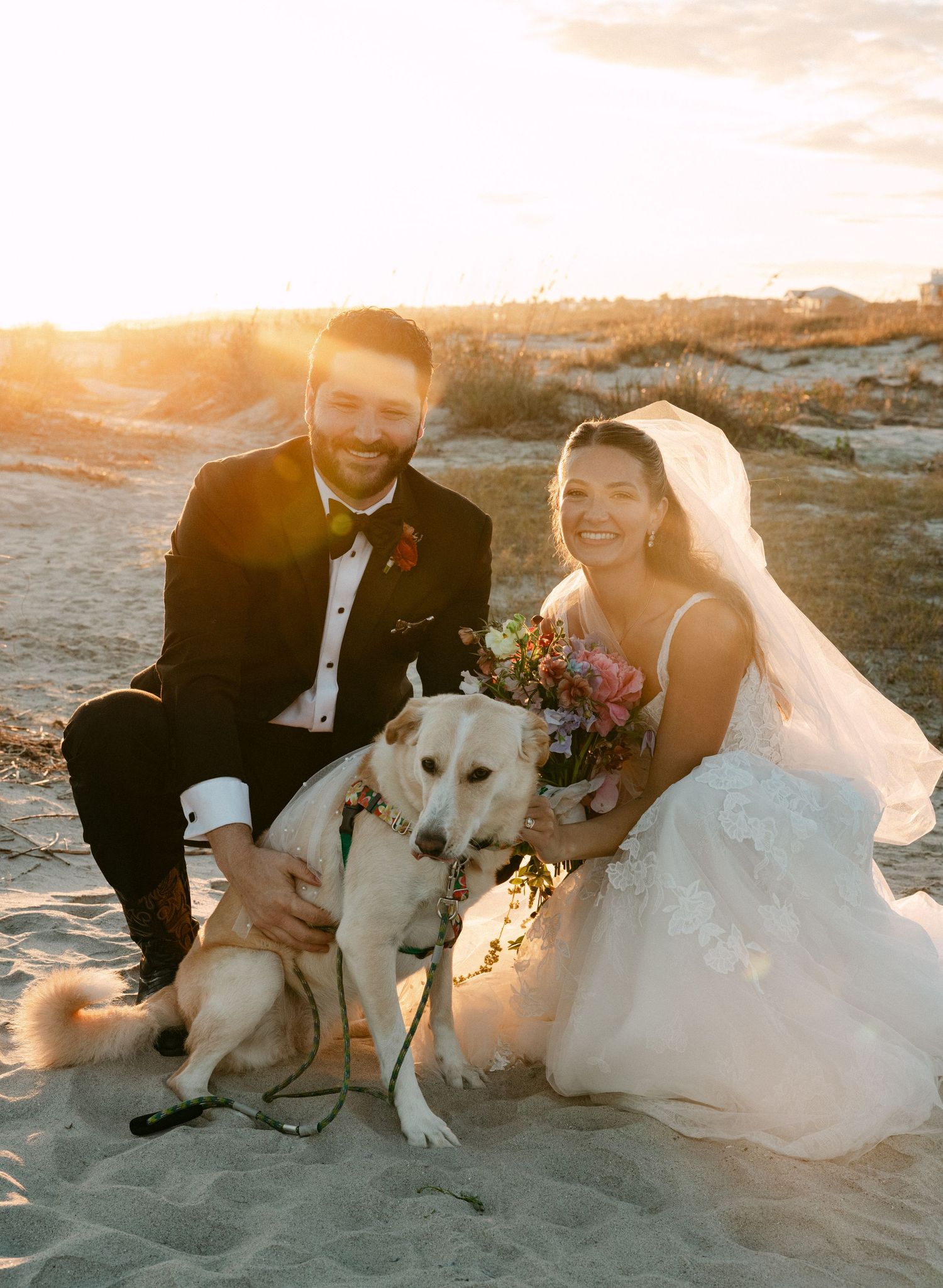 Bride and groom pose with golden Labrador on sandy beach at sunset, bride holding colorful bouquet wearing veil.