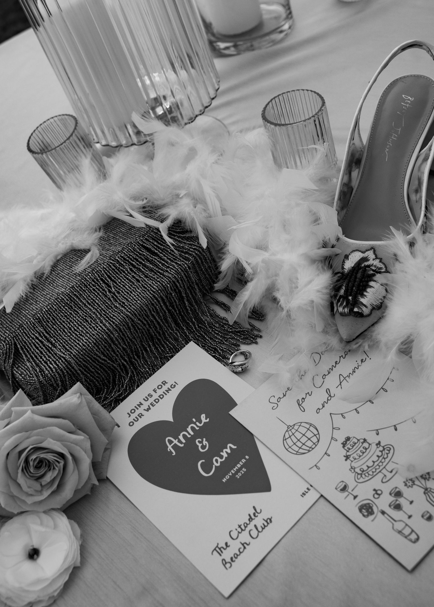 Black and white flat lay of Annie and Cam wedding invitation, feather boa, clutch bag, heels, and floral decor.