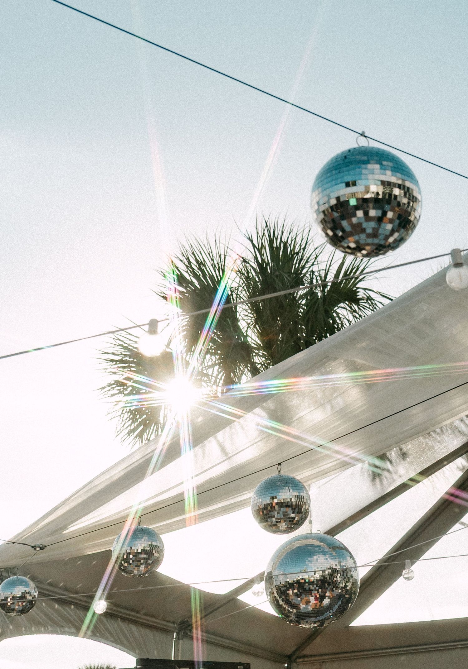 Sparkling disco balls hanging under a tent canopy with sunlight flares and tropical palm trees.