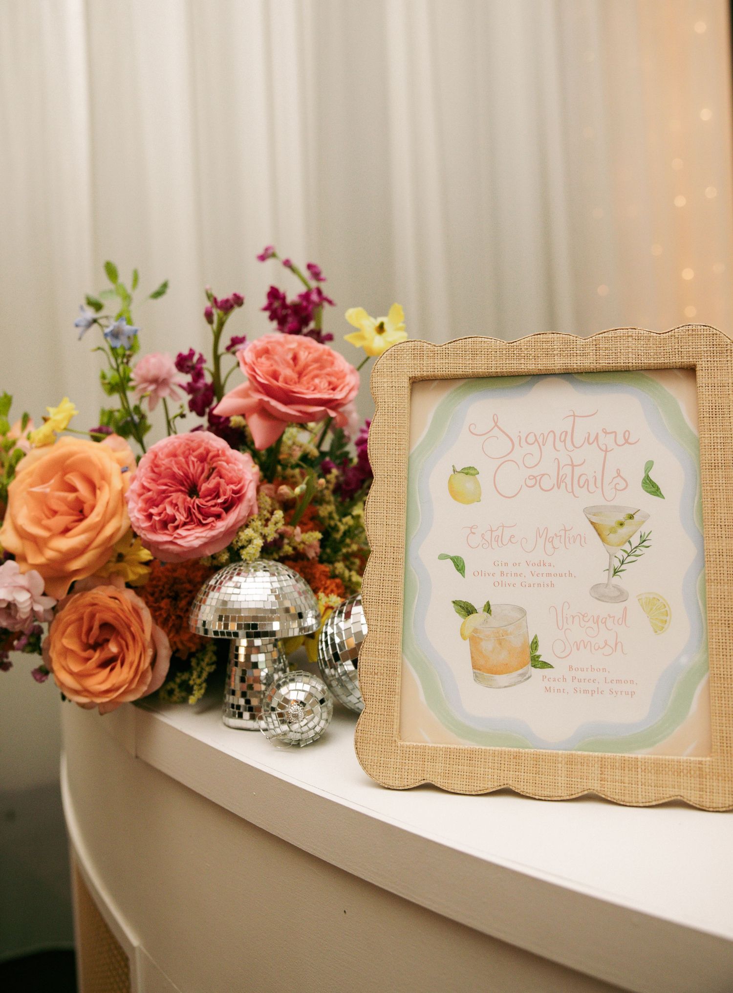 Framed signature cocktails menu beside colorful floral arrangement with disco balls on white table at wedding reception.