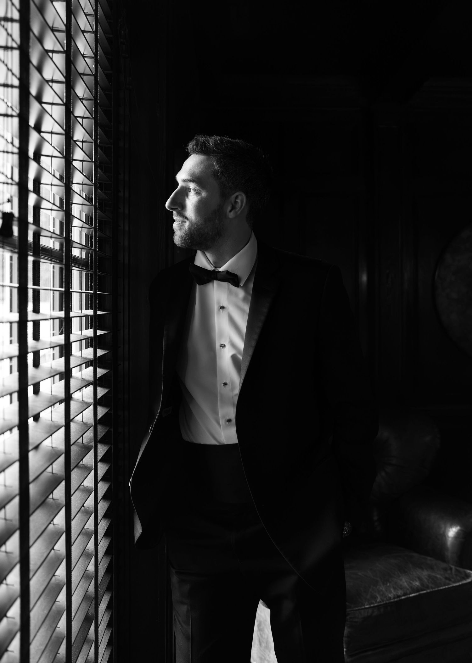 Groom in black tuxedo and bow tie gazes out window in dramatic black and white portrait.
