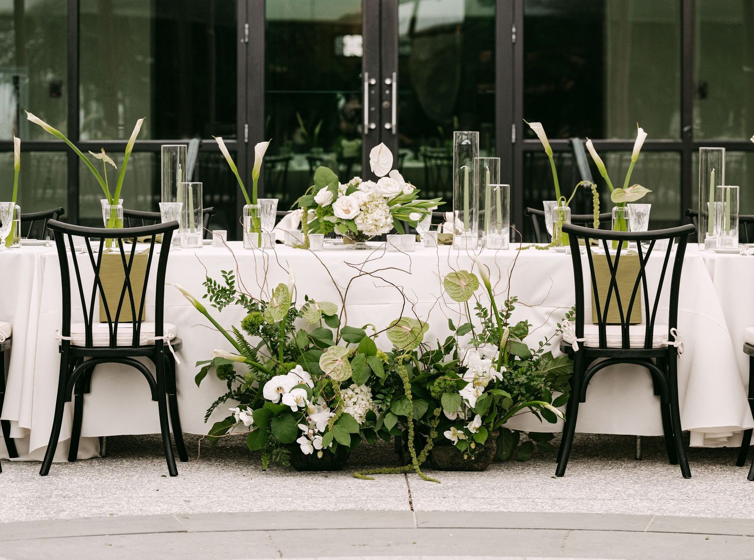Elegant wedding sweetheart table with white floral arrangements, greenery, and black chairs.