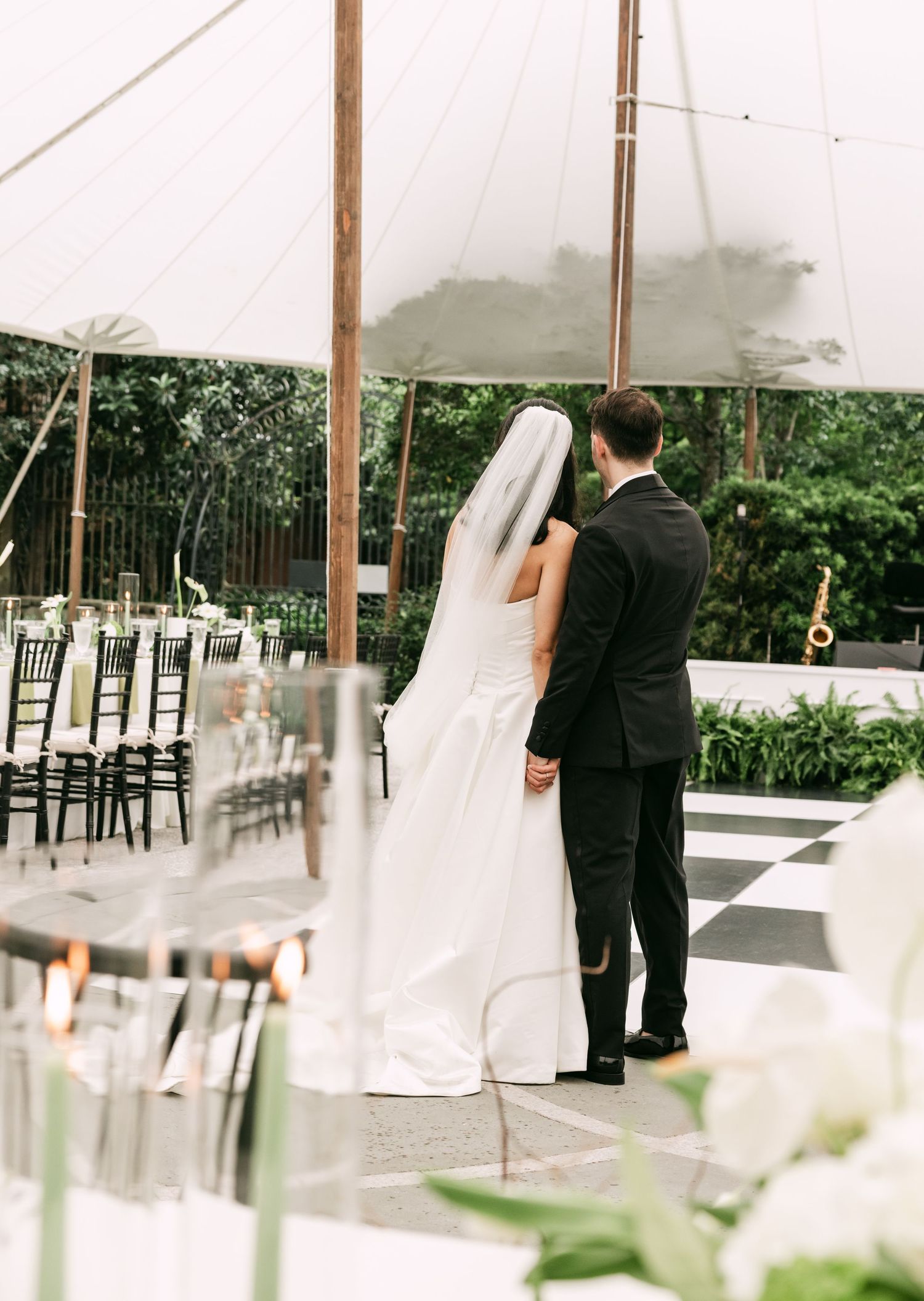 Bride and groom holding hands under a white tent overlooking a decorated reception with checkered dance floor.