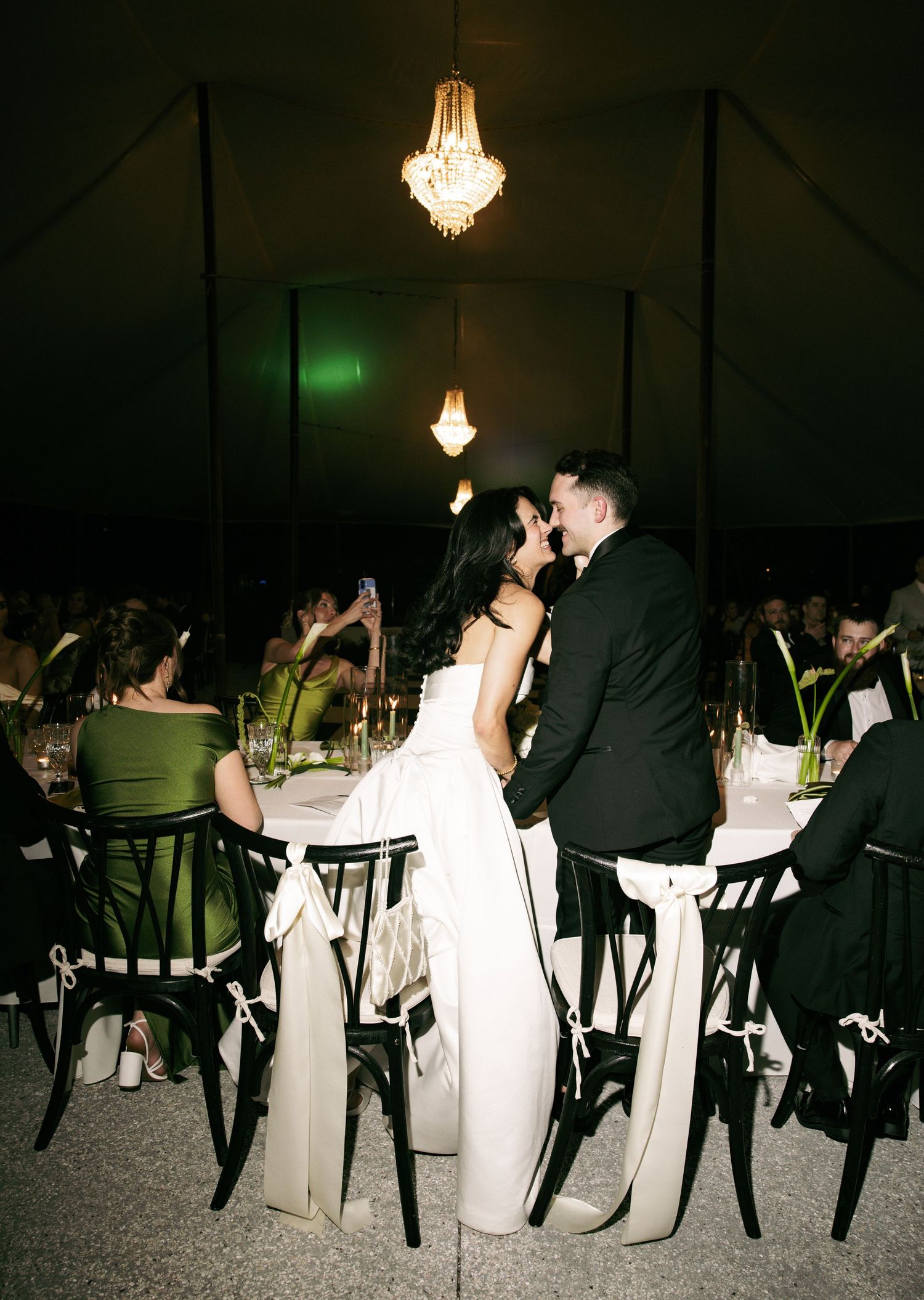 Bride and groom share a kiss at their elegant wedding reception under chandelier lighting in a dark romantic tent venue.