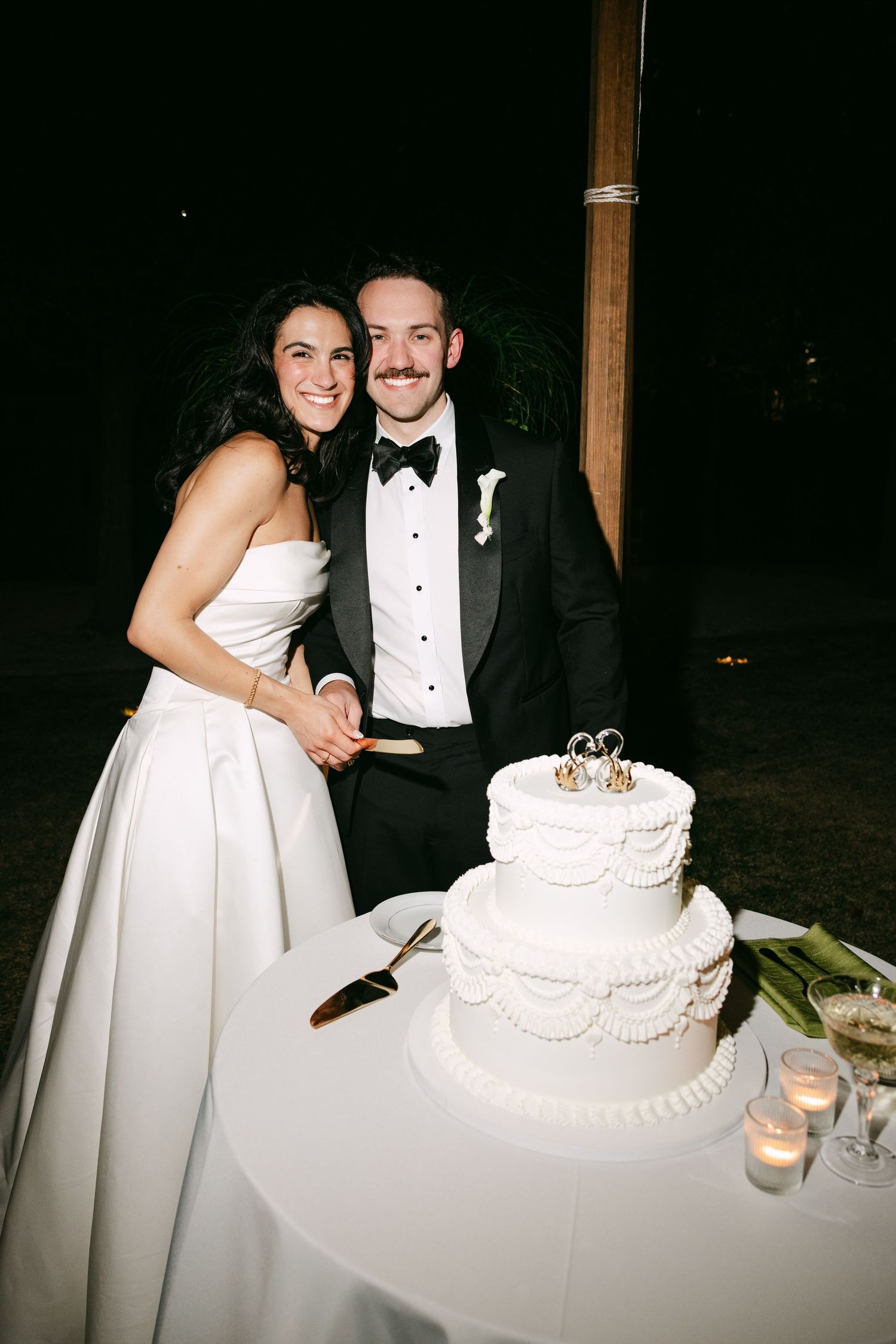 Bride and groom smiling while cutting their two-tier white wedding cake decorated with crown toppers at night.