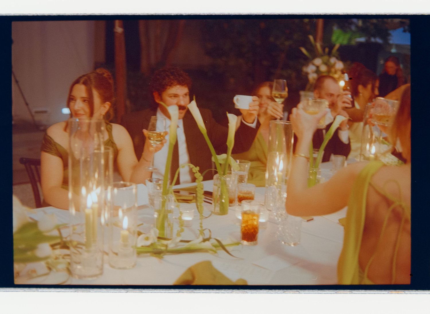 Wedding guests raising glasses for a toast at candlelit dinner table with calla lily floral arrangements.