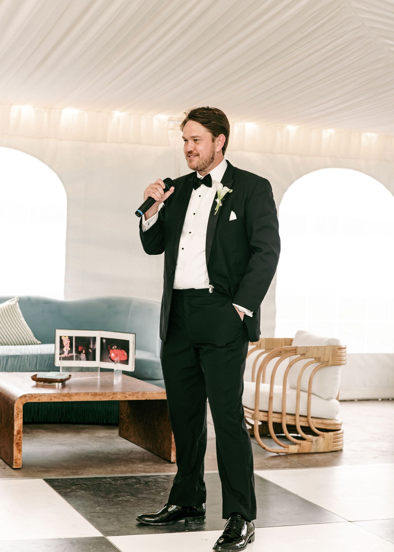 Groom in black tuxedo with bow tie speaks into microphone during wedding reception speech inside white tent.