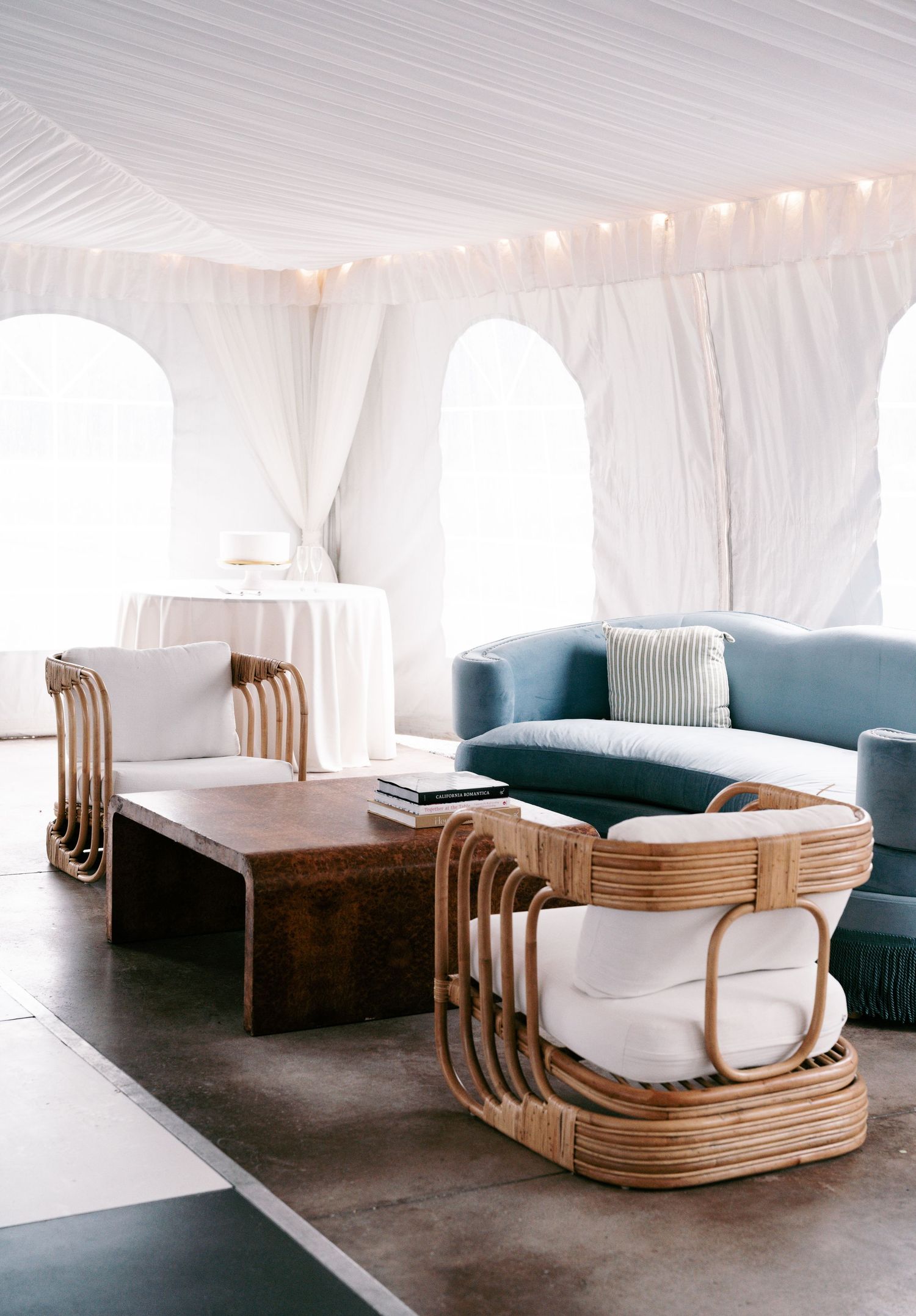 Elegant wedding tent lounge with rattan chairs, blue velvet sofa, wood coffee table, and white draped ceiling lights.