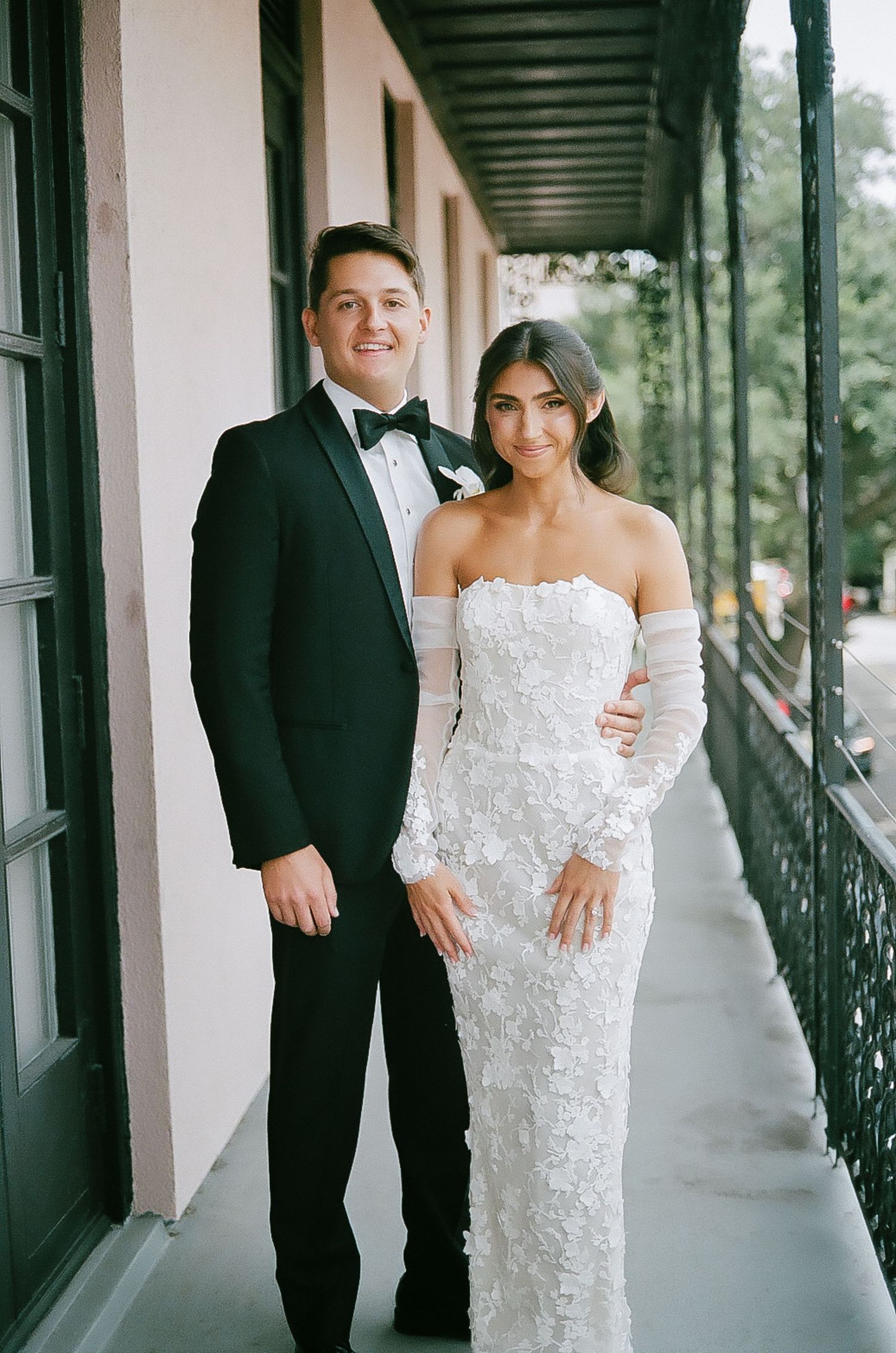 Elegant couple poses in wedding attire on a covered outdoor walkway with white columns.