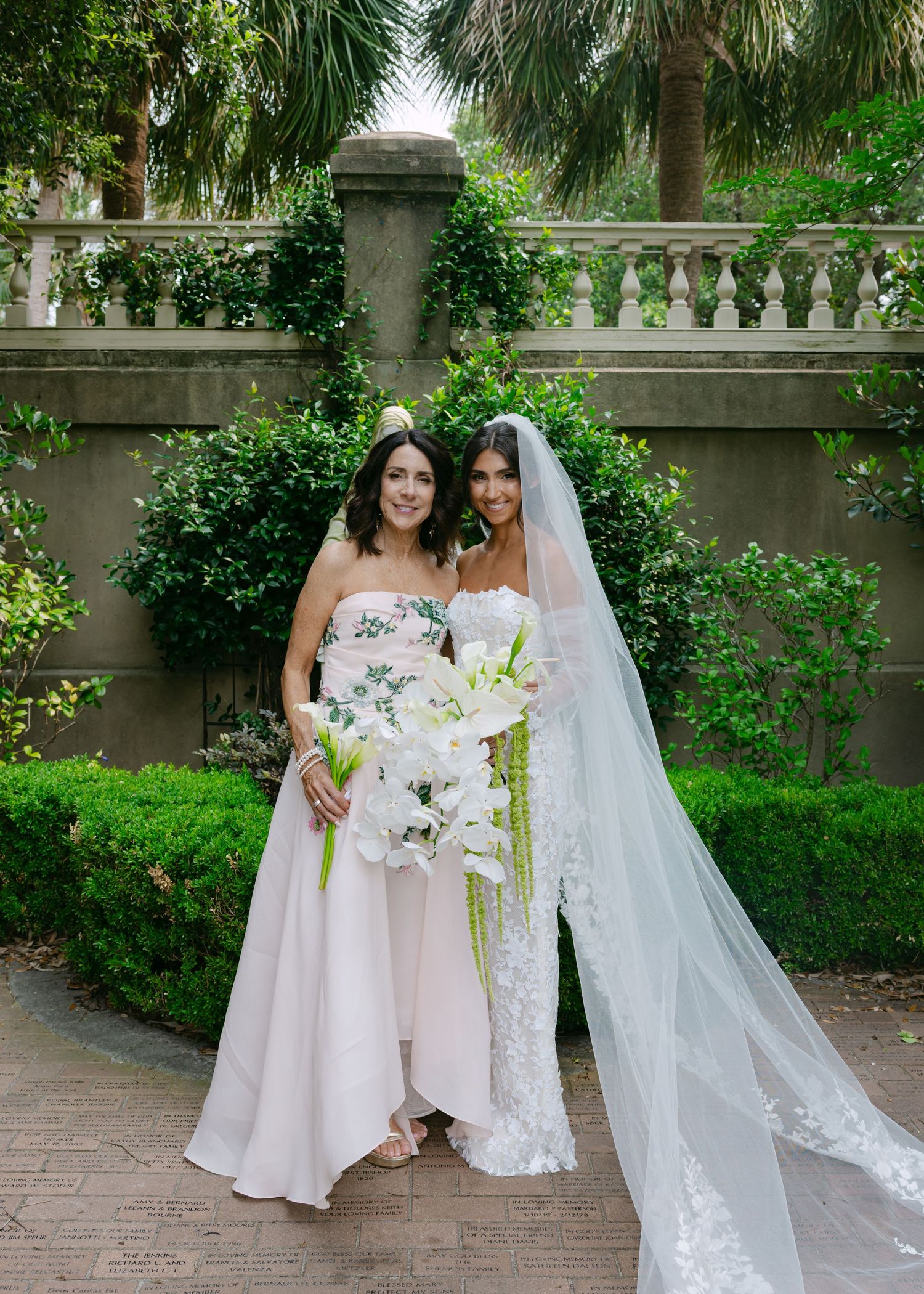 Elegant bridal party with flowing dresses and white floral bouquets pose in garden setting.