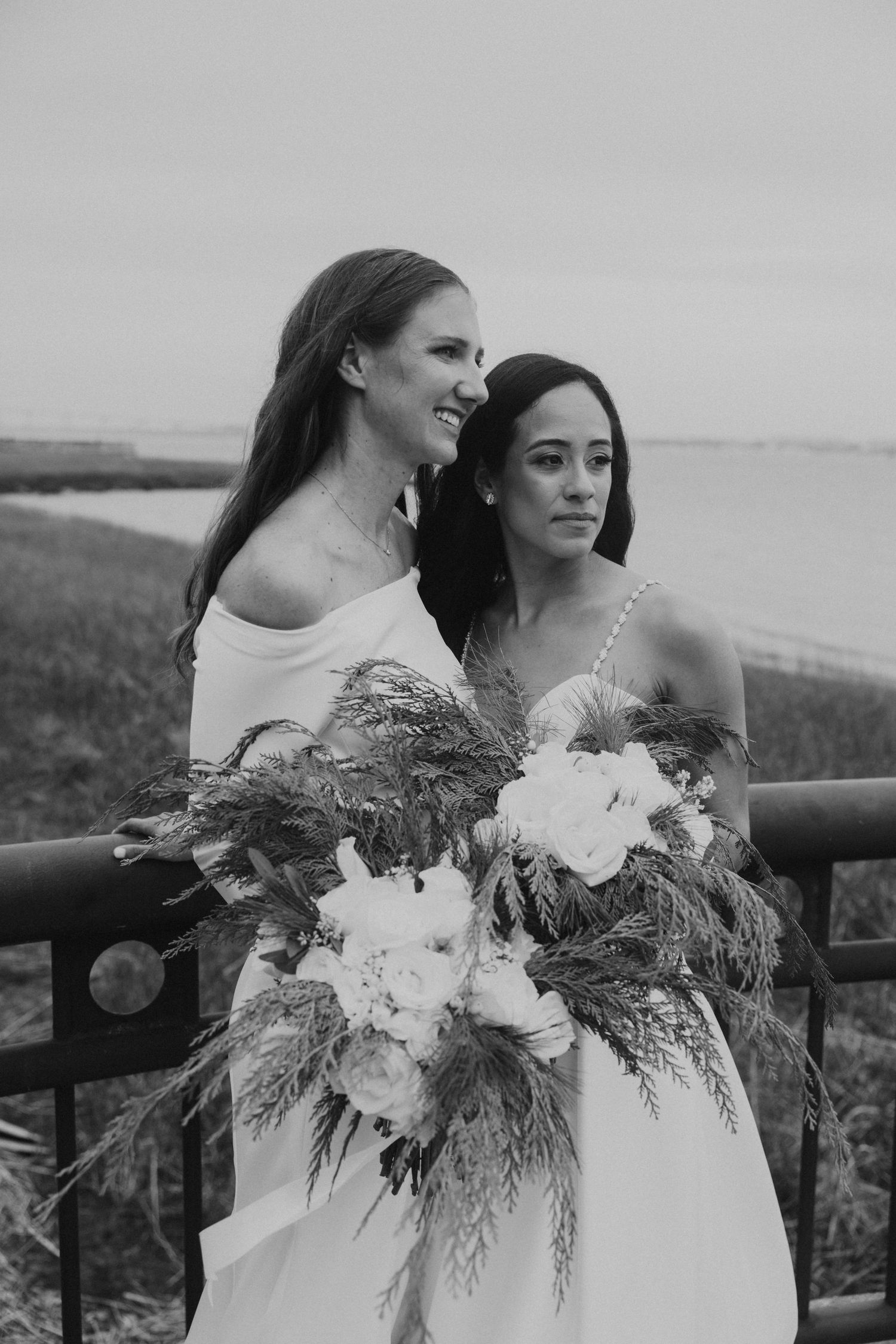 A couple in white dresses holds elegant floral bouquets against a misty coastal backdrop.