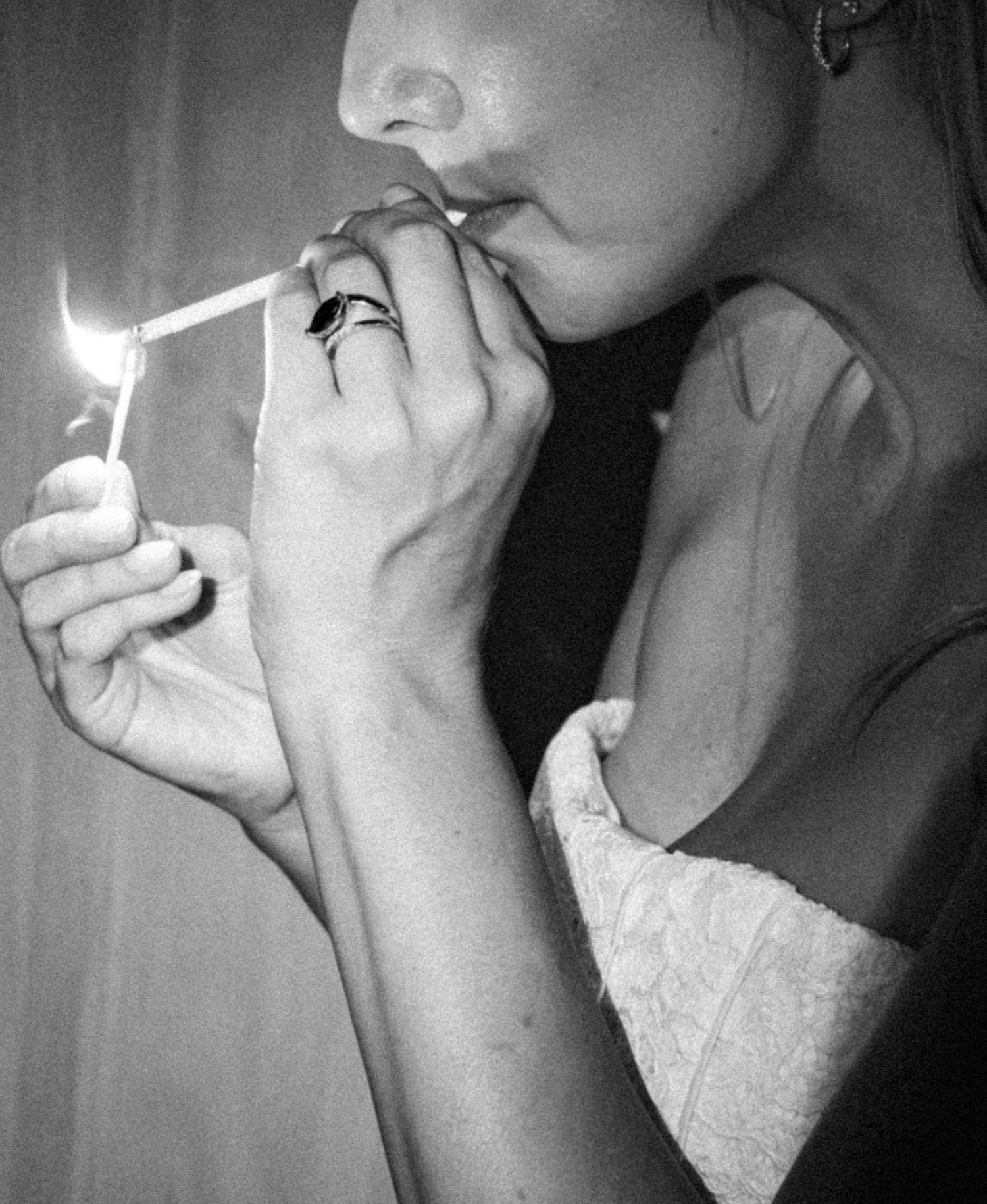 Black and white photo of a woman lighting a cigarette with a lighter, wearing rings on her fingers.