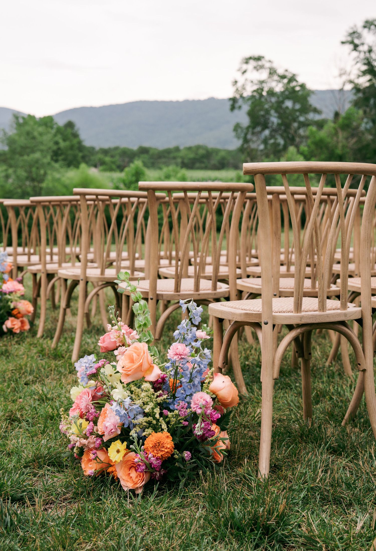Colorful floral arrangements with pink, orange, and blue blooms line wooden chairs at an elegant outdoor wedding ceremony.