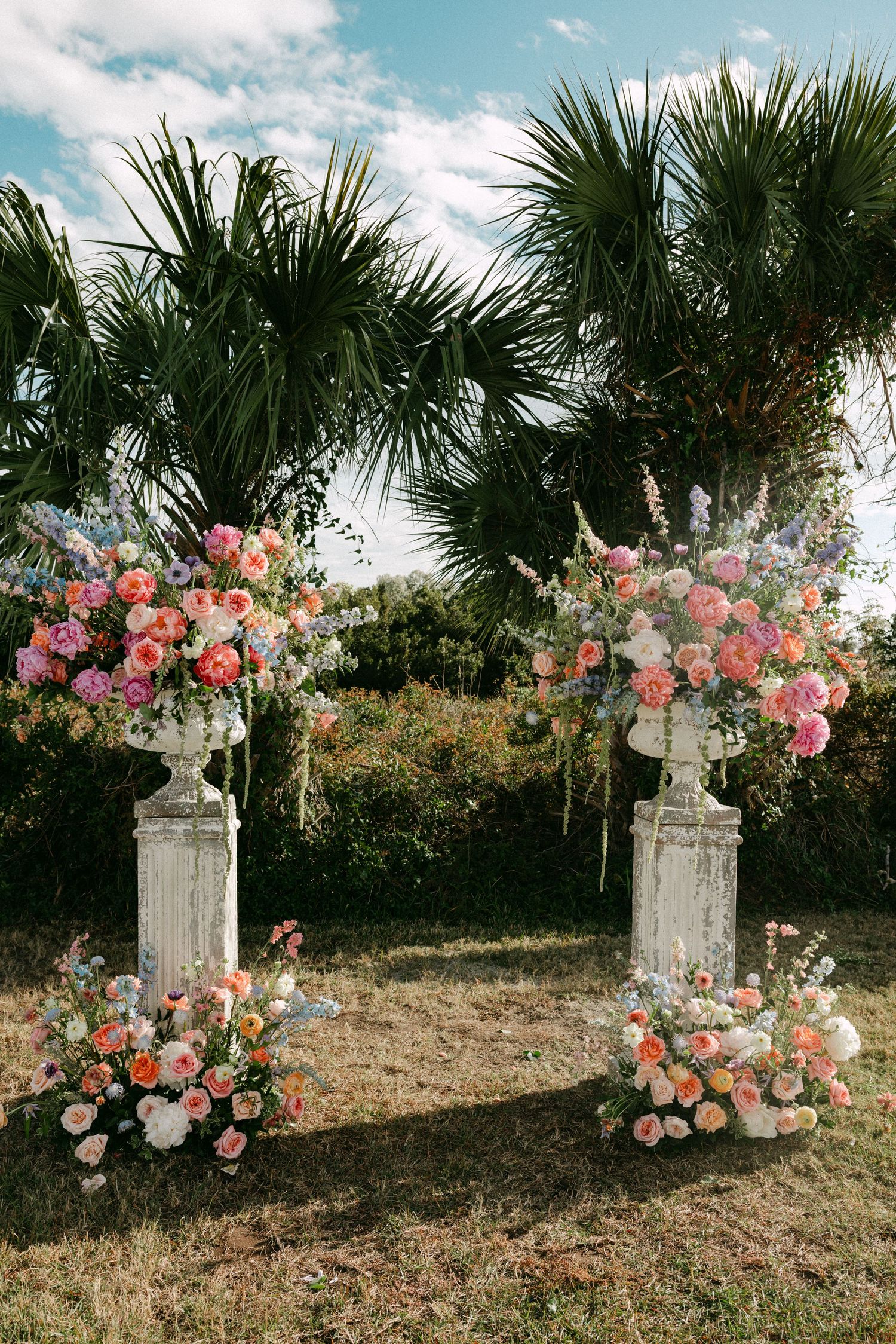 Outdoor wedding ceremony arch with colorful pink, coral, and blue floral arrangements on white stone pedestals near palm trees.