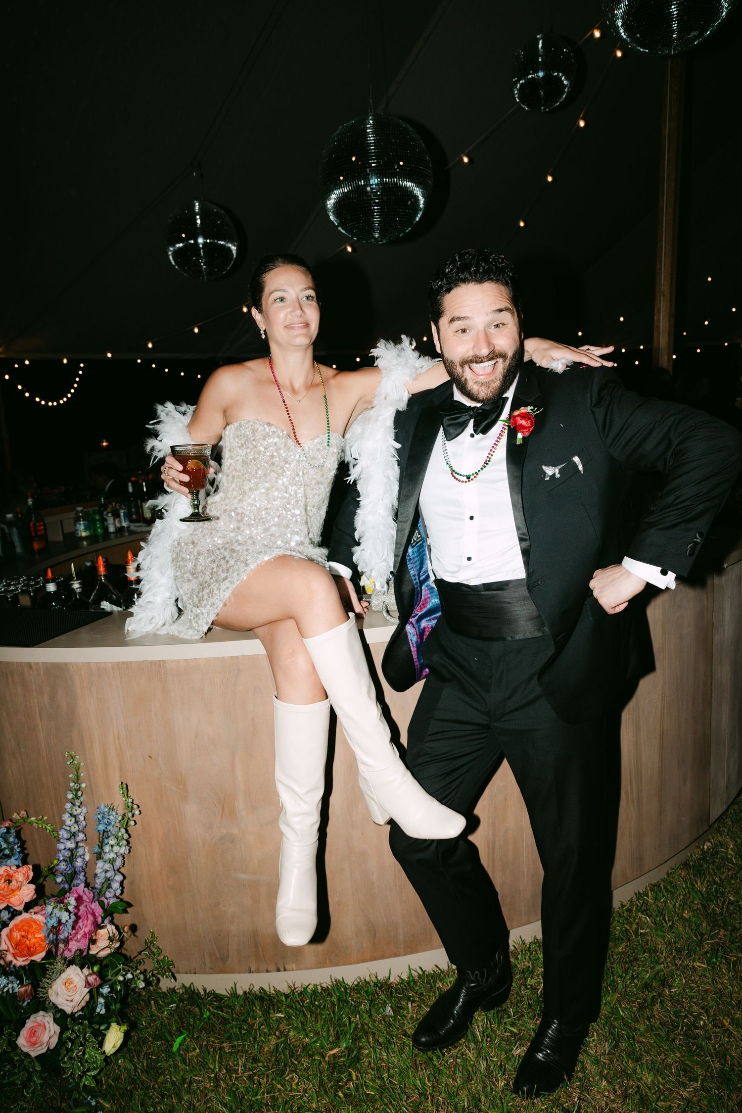 Bride in a sequined dress with white feather boa and knee-high boots poses with groom in tuxedo at wedding reception bar.