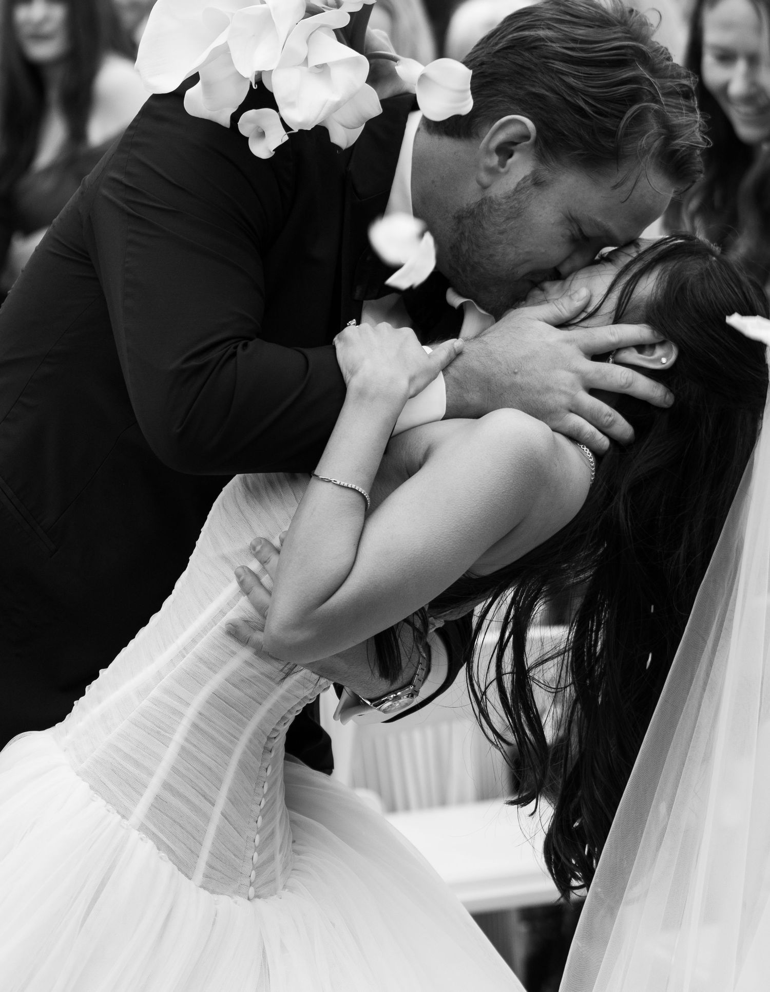 Black and white wedding photo of a couple sharing a romantic dip kiss, bride in corset gown with flowers in hair.