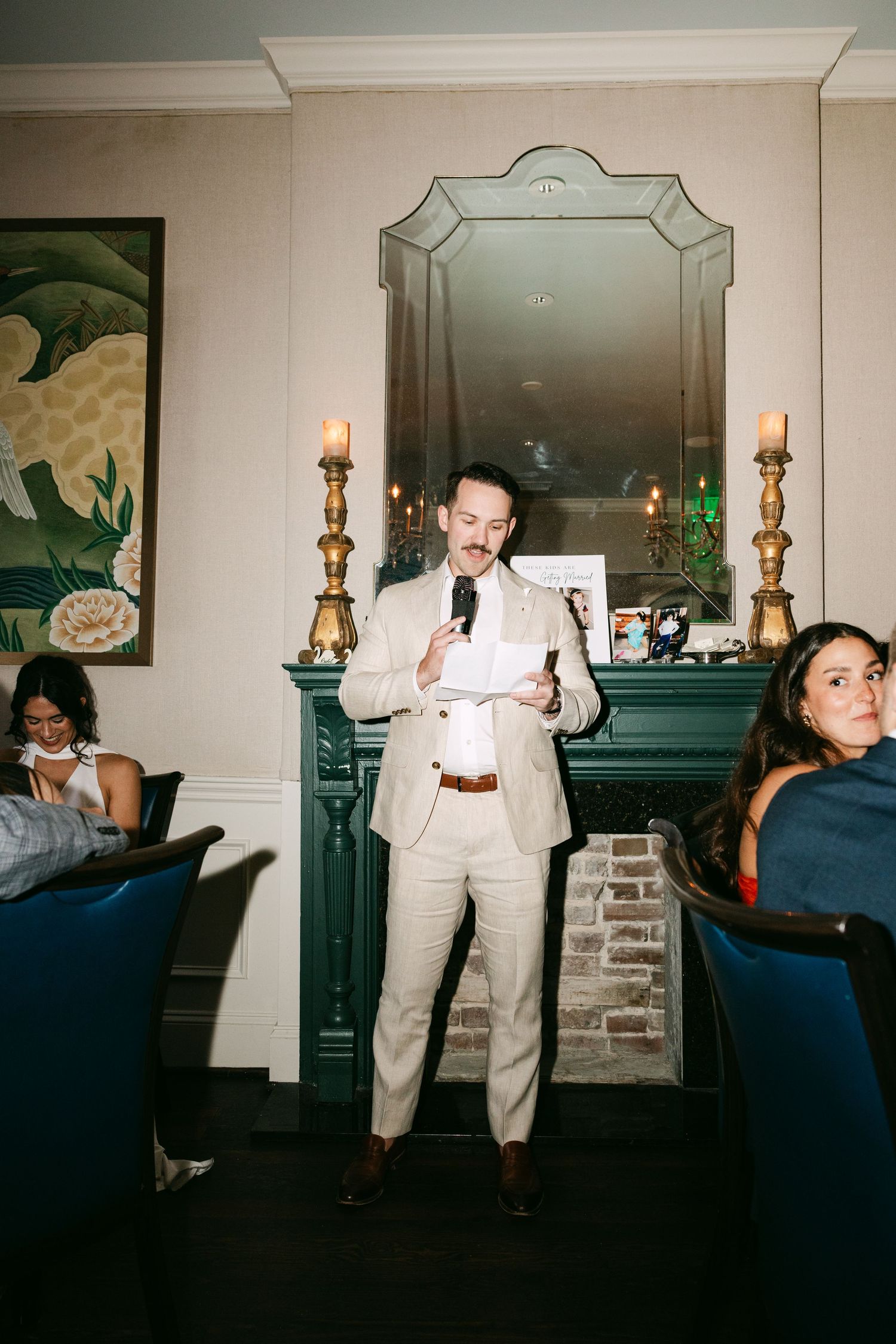 Man in beige suit giving a speech at a wedding rehearsal dinner beside a green fireplace.