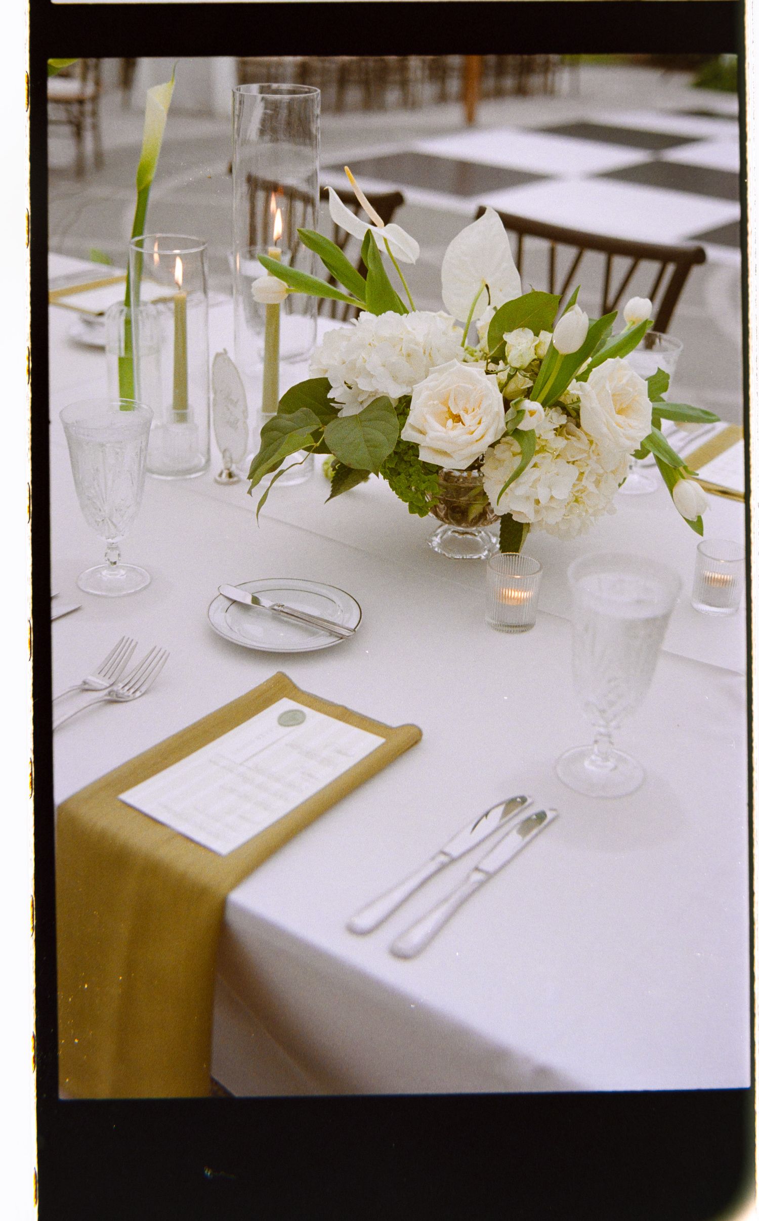 Elegant wedding reception table with white floral centerpiece, gold napkin, crystal glassware, and candles on white linen.