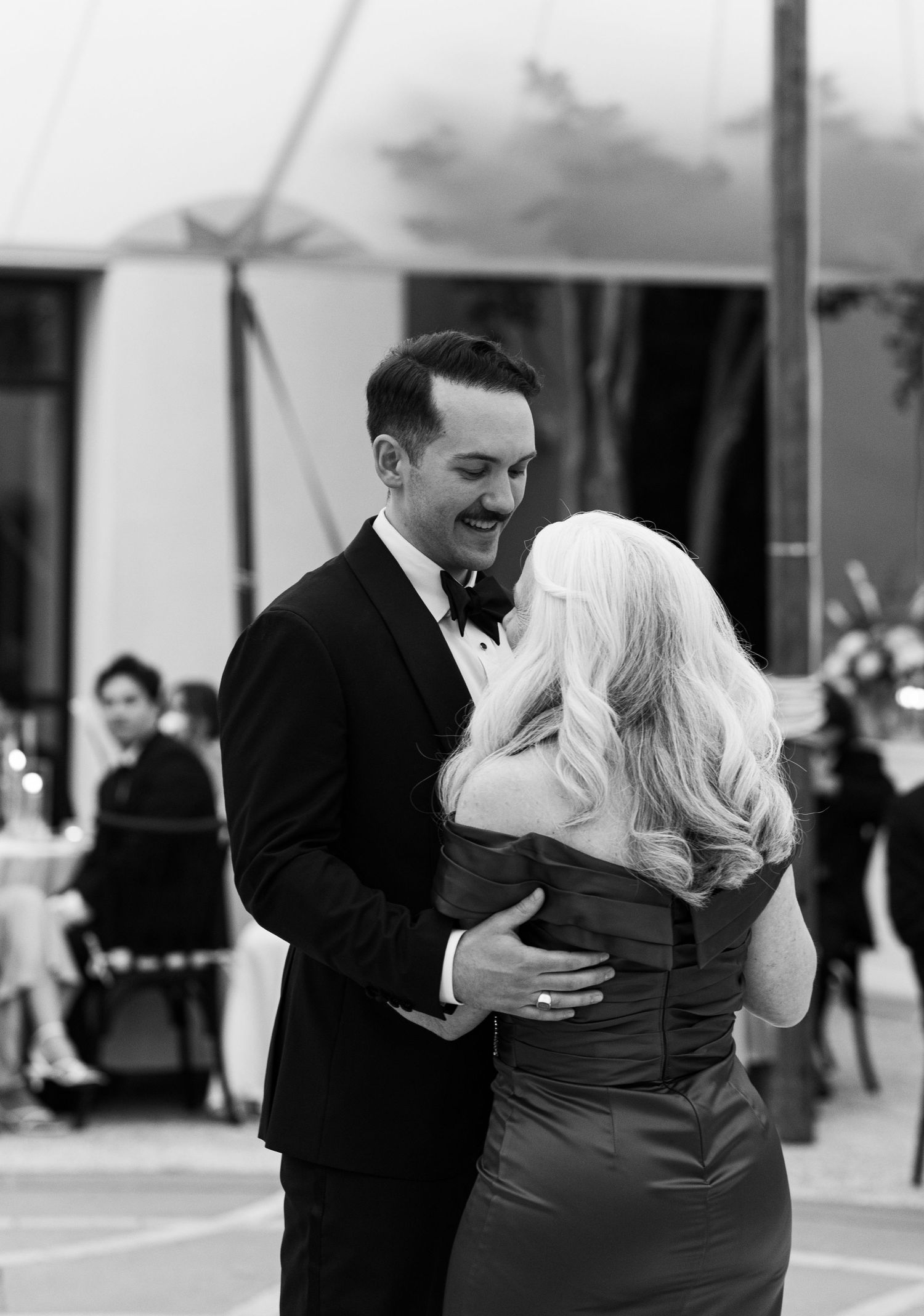 Black and white photo of groom in tuxedo sharing a tender mother-son dance at outdoor wedding reception.