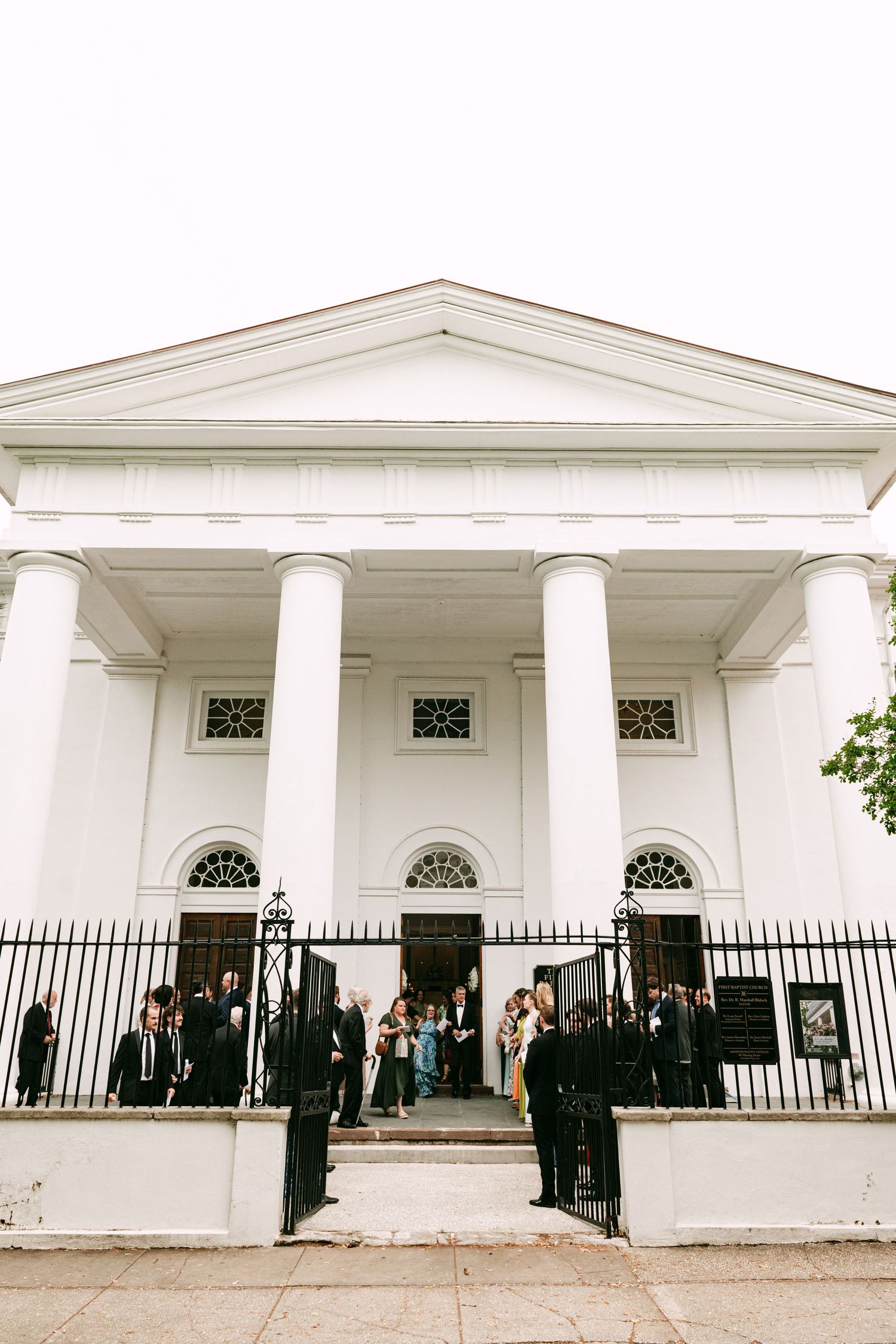 Wedding guests gathered outside a white neoclassical church with large columns and black iron gates.