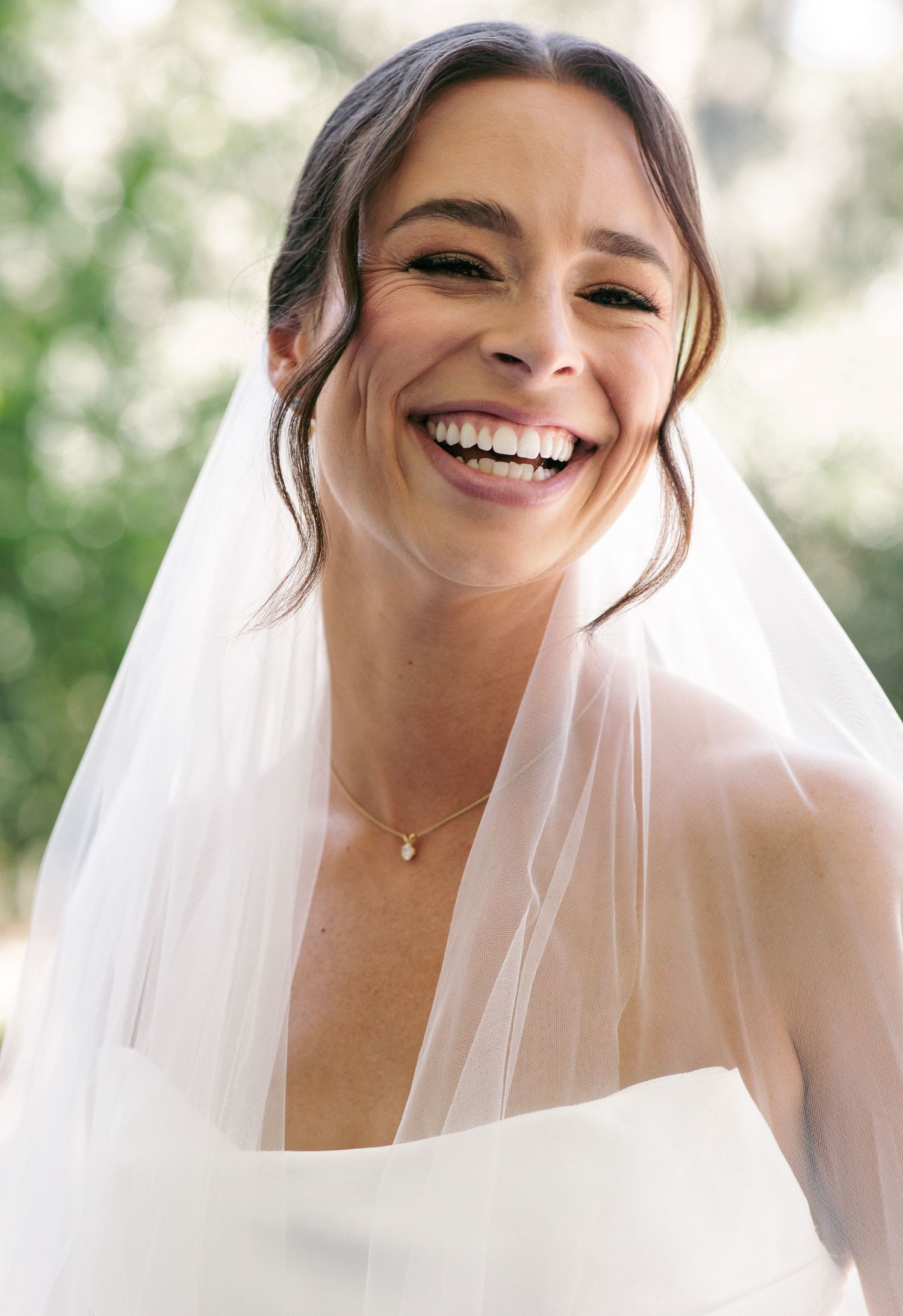 A radiant bride smiles joyfully wearing a white wedding dress, delicate veil, and a simple pendant necklace outdoors.