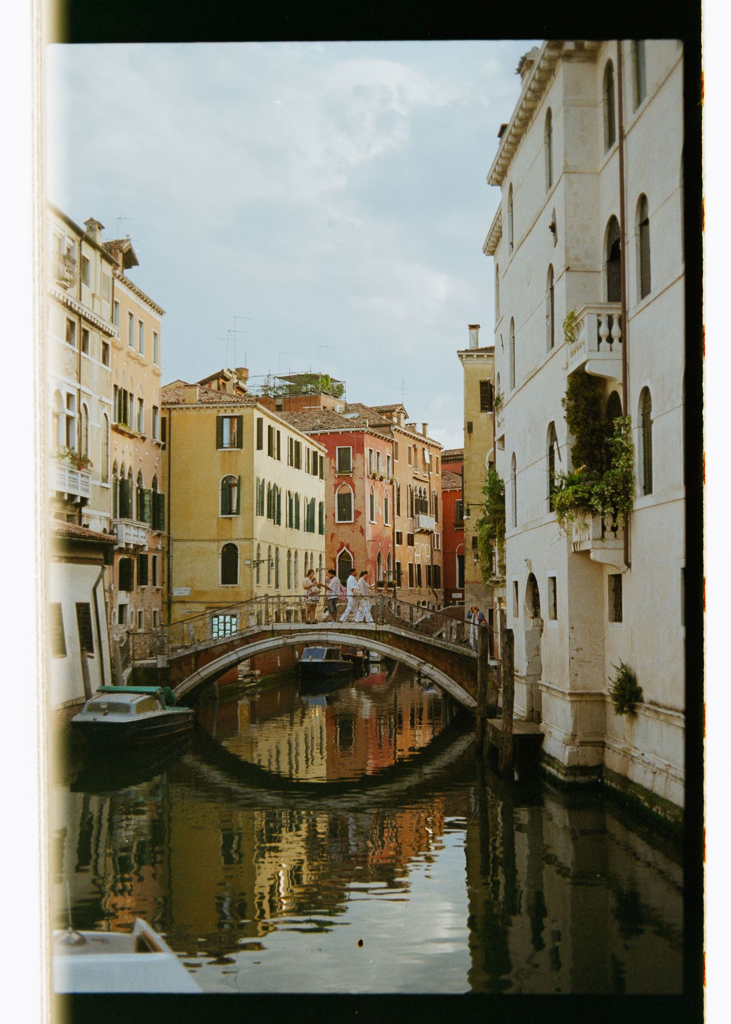 A picturesque Venice canal with a stone bridge, colorful historic buildings, and calm reflective waters under a cloudy sky.