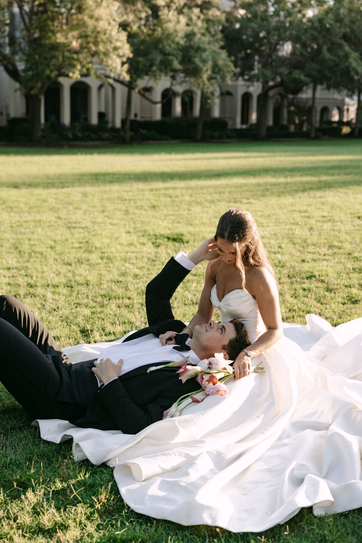 Bride in white gown and groom in black tuxedo lying on green grass in romantic wedding portrait outdoors.