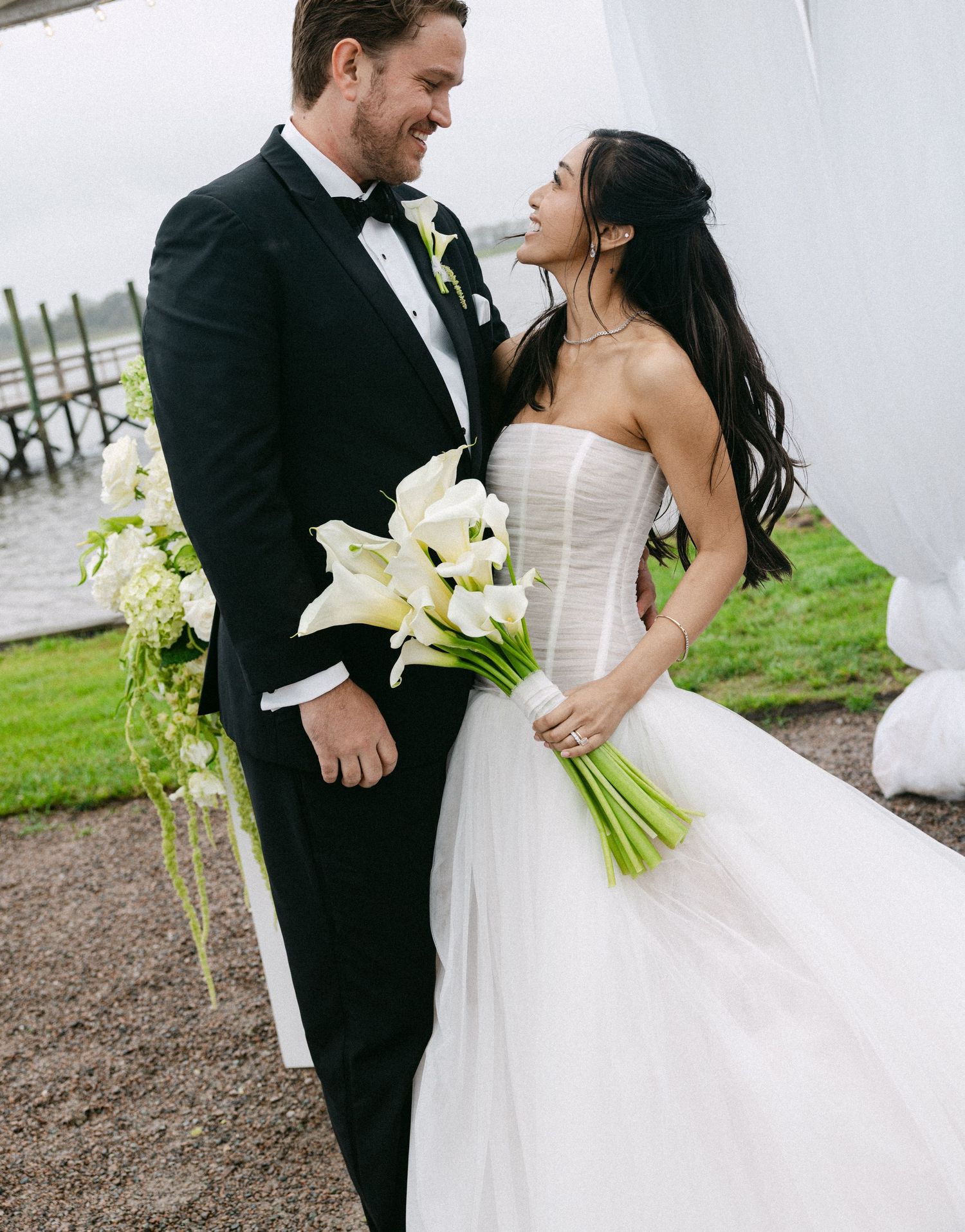 Bride in strapless white gown holding calla lily bouquet smiles at groom in black tuxedo at outdoor waterfront wedding.