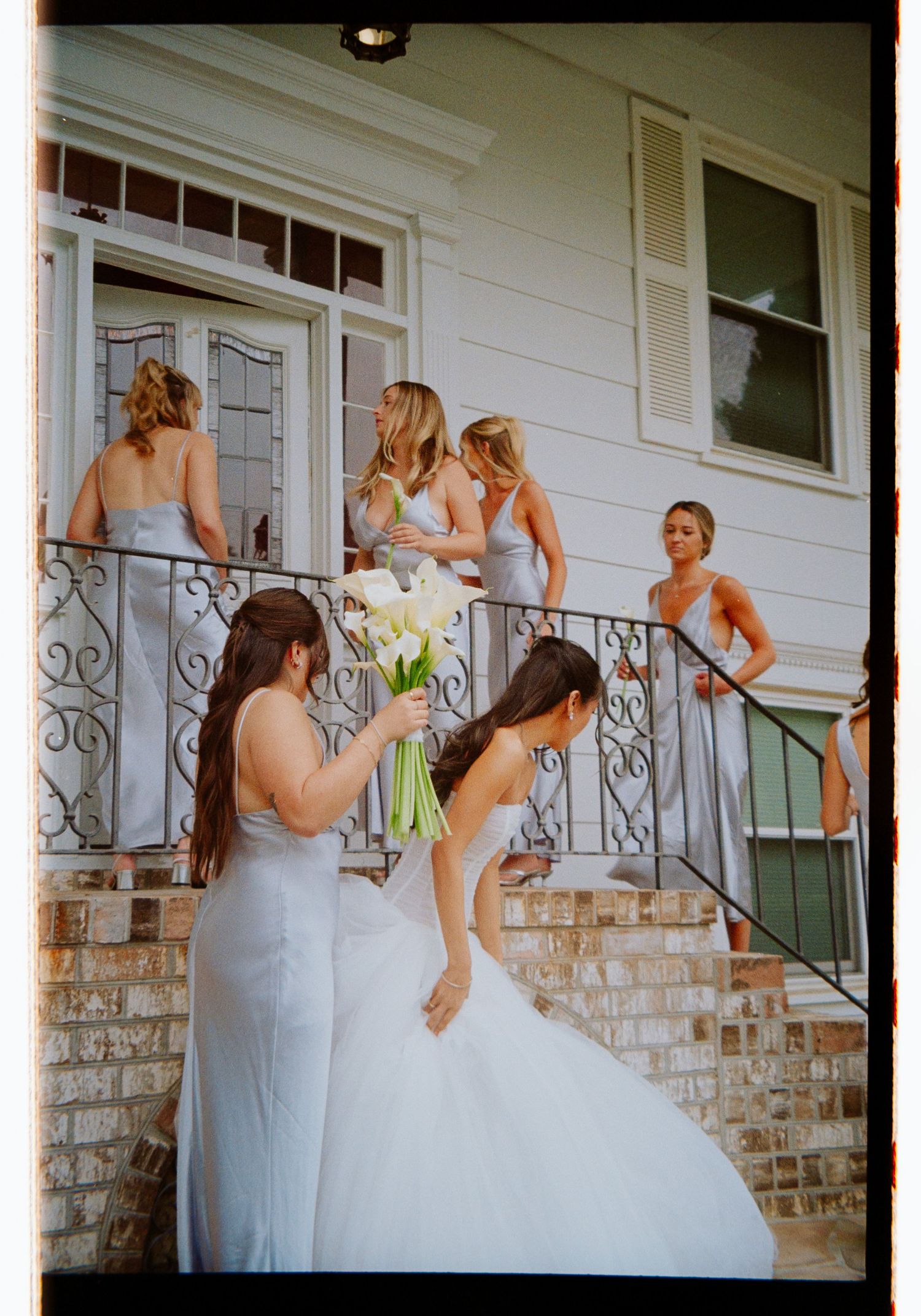 Bride in white gown holds calla lily bouquet surrounded by bridesmaids in silver dresses on brick staircase.