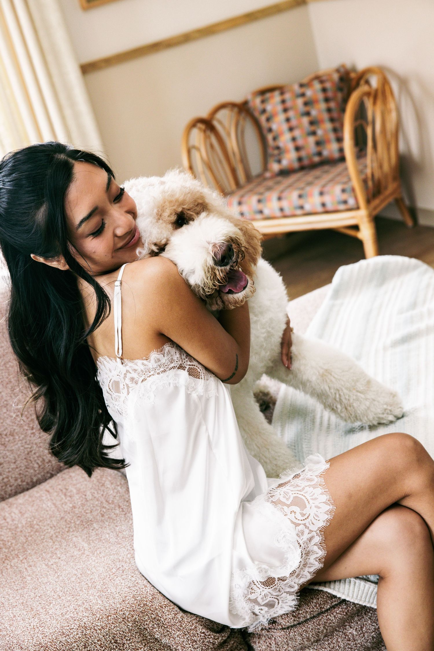 Bride in white lace slip dress hugging her fluffy golden doodle dog on a couch before her wedding.